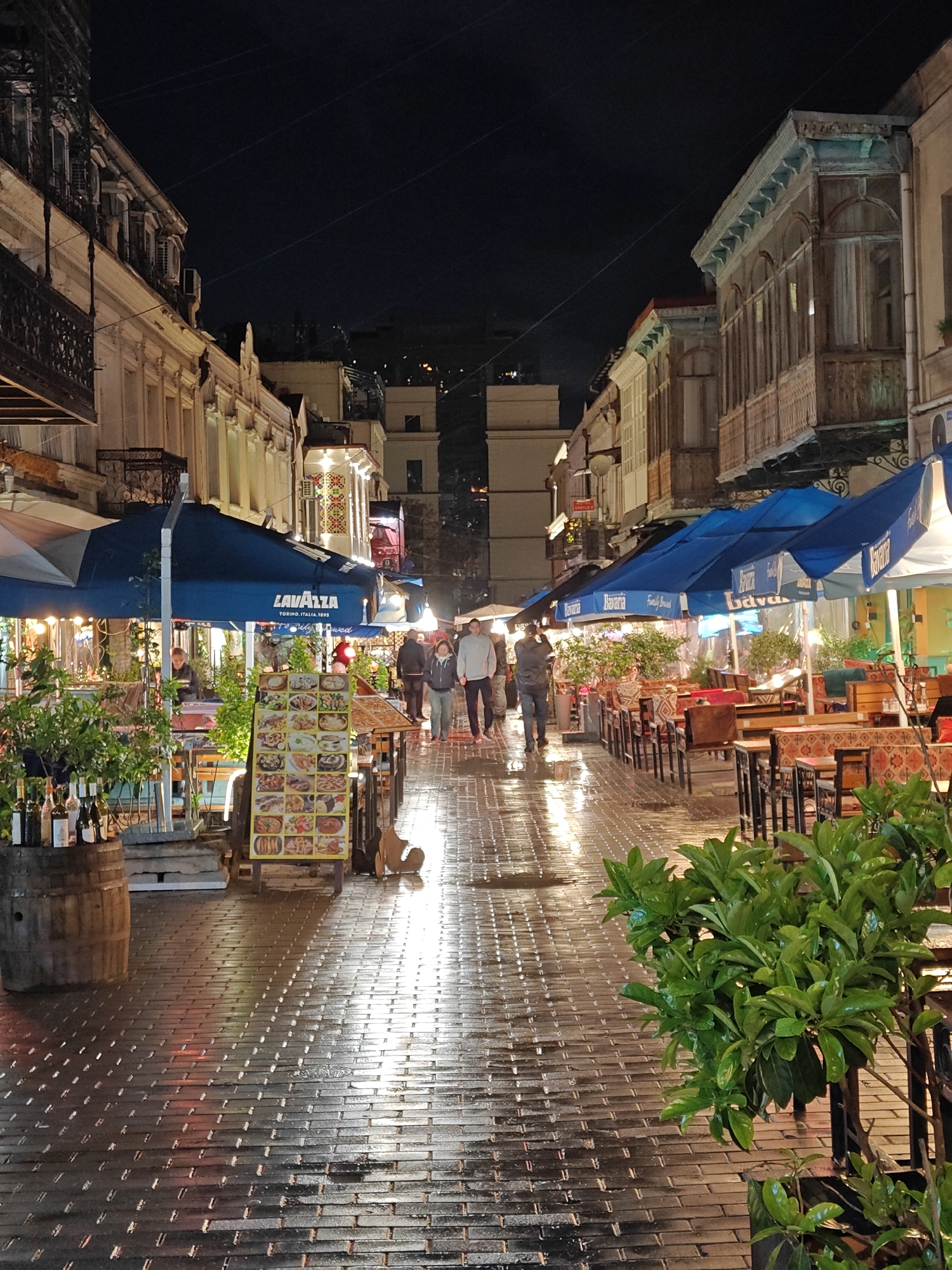This nighttime photograph depicts a charming, wet, cobbled street in Tbilisi, Georgia, illuminated by artificial lights after a recent rain. The wet pavement creates striking reflections of the surrounding lights, forming a radiant path through the scene.
The street is lined with multi-story buildings showcasing traditional Georgian architecture, including ornate balconies, some of which are enclosed. Numerous cafes and restaurants occupy the ground floors, extending their outdoor seating areas onto the street under large blue umbrellas. Visible branding on these umbrellas includes "LAVAZZA TORINO, ITALIA, 1895" on the left and "Bavaria" on the right.
Several people are seen strolling along the street in the middle distance, appearing to enjoy the evening. Tables and chairs, some adorned with patterned tablecloths, are set out for diners. On the left, a wooden barrel holding bottles, likely wine, stands near a prominent upright menu board displaying various food items. Potted green plants are used as decorative elements. The dark night sky contrasts with the brightly lit street, creating a lively yet peaceful ambiance characteristic of a historic district.