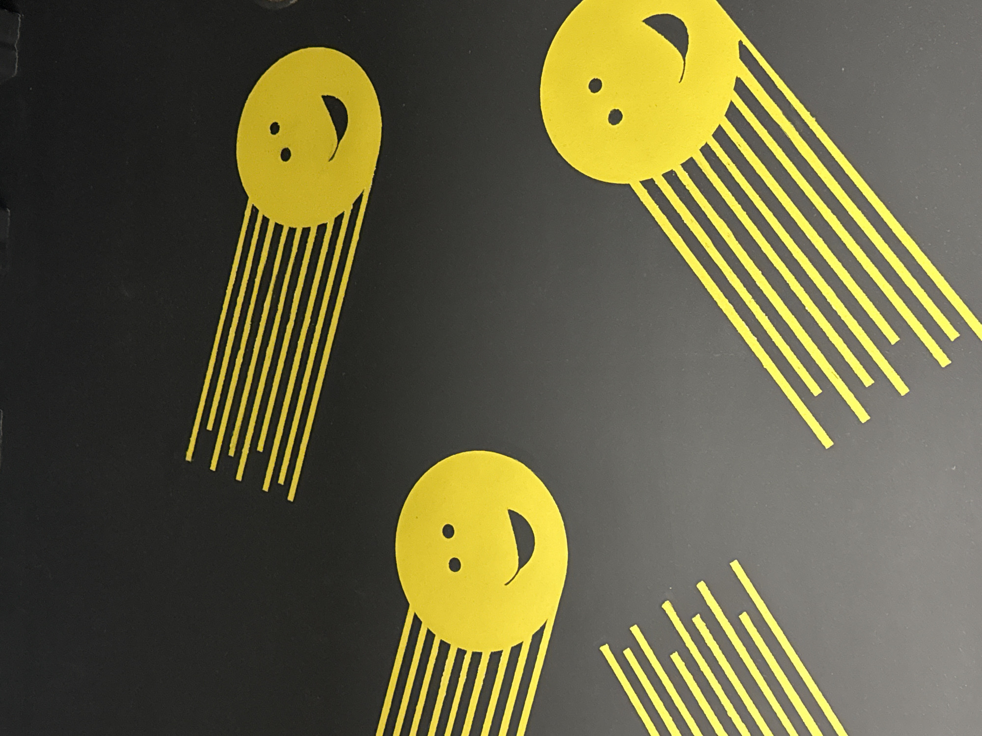 The image shows a close-up of a dark grey or black surface with three yellow graphic elements. Each element consists of a yellow circle with two dots for eyes and a semi-circular mouth, resembling a smiley face. From each of these "smiley faces" extends a series of parallel yellow lines, resembling a comet or shooting star. The faces are oriented in different directions, with one in the upper left, one in the upper right, and one in the lower center of the frame. The lines extending from the faces also vary in length. There is no indication of the location in Votkinsk, Russia.