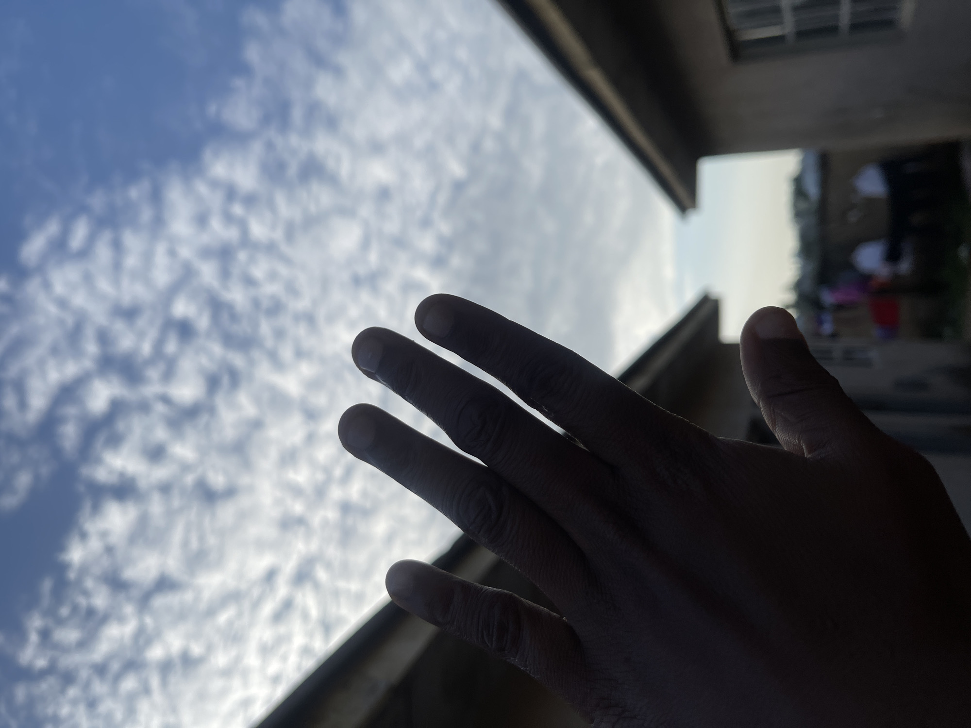 A person's hand, silhouetted against a cloudy blue sky, is held up in the foreground. The hand obscures the upper portion of the image, with fingers splayed. In the background, between two buildings, a blurred scene depicts a courtyard with several individuals. The buildings appear to be simple residential structures, suggesting a locale in Jos, Nigeria. The overall impression is of an outdoor scene, possibly in the afternoon.