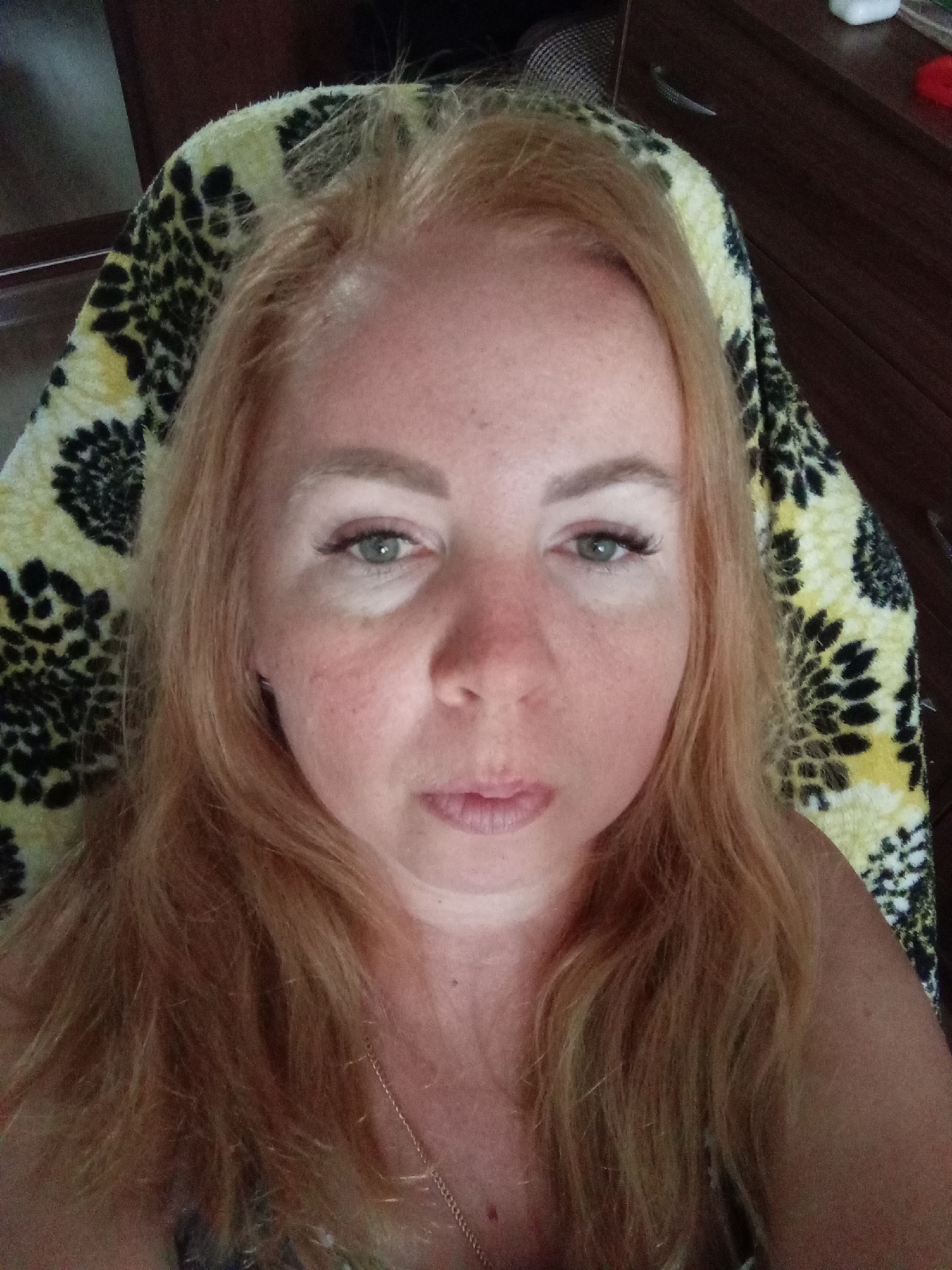 The image is a medium shot of a Caucasian woman with blonde hair, looking directly at the camera. She appears to be indoors, with a patterned fabric, possibly a blanket or cushion, behind her head and shoulders. The fabric has a black and yellow floral or abstract pattern. To the right of the woman, a dark brown wooden piece of furniture, likely a dresser or cabinet, is visible. The lighting suggests it is daytime, possibly natural light from a window. There is no indication of the specific location within the city of Roshal, Russia.