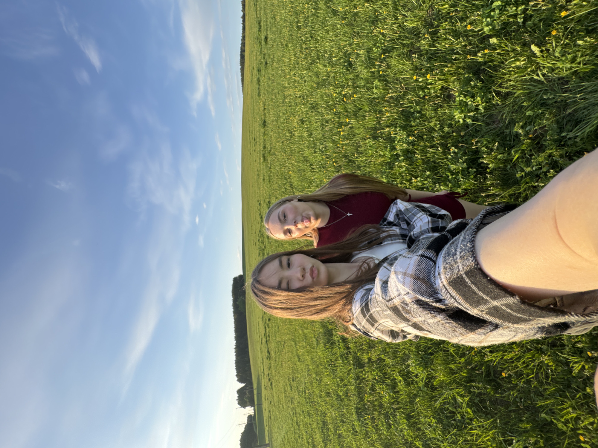 Two young women lie side-by-side in a grassy field, likely located in the countryside of Июльское, Russia. The sky above is blue with scattered clouds. The field extends into the distance, bordered by a treeline. The woman closer to the camera wears a black and white plaid shirt. The second woman wears a maroon shirt and has her tongue sticking out playfully. Both are positioned on their backs within the grass.