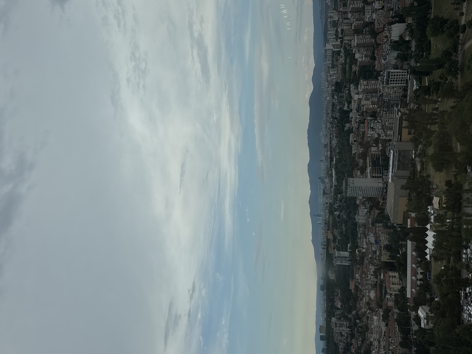 The image depicts a panoramic view of Sofia, Bulgaria, showcasing a sprawling cityscape under a cloudy sky. In the foreground, a section of a park is visible, characterized by lush green trees and a cluster of white tents, possibly indicating an event or market. Adjacent to the park, a parking lot filled with numerous cars is present.
As the view extends into the city, a dense urban landscape unfolds, with a variety of buildings ranging from low-rise residential structures with red-tiled roofs to taller, modern office buildings and apartment complexes. A notable structure in the mid-ground appears to be a large, rectangular building with a light-colored facade, possibly a commercial or public facility. In the background, a mountain range forms a natural backdrop to the city, its peaks softened by distance and the atmospheric haze. The sky is predominantly overcast with varying shades of grey and white clouds, with hints of blue peeking through in some areas, suggesting a subdued lighting condition. The overall impression is that of a typical urban environment on a cloudy day.