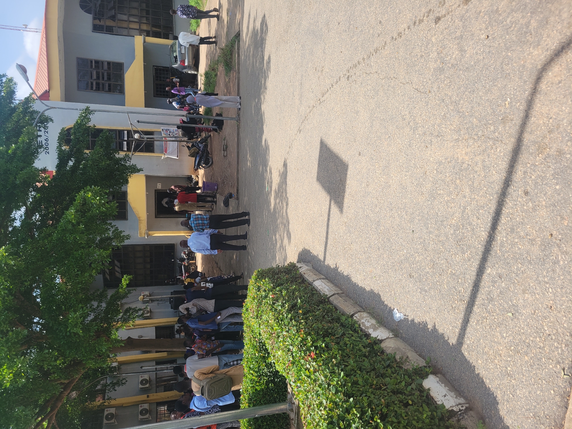 The image depicts the exterior of a building, likely an educational institution, in Ogbomosho, Nigeria. The building has a light gray facade with yellow trim and prominent windows. A large tree with lush green foliage shades a portion of the building and the surrounding area. Several individuals, predominantly young men dressed in casual attire, are gathered outside the building. Some are standing in groups, engaged in conversation, while others appear to be waiting. Motorcycles and a silver sedan are parked near the entrance. The ground is paved with asphalt, and a neatly trimmed hedge runs along the left side of the image. The scene is illuminated by natural sunlight, casting shadows on the ground. A sign on the building displays text, partially obscured by the tree, including "2006/200". The overall atmosphere appears to be one of informal gathering or activity outside the building.