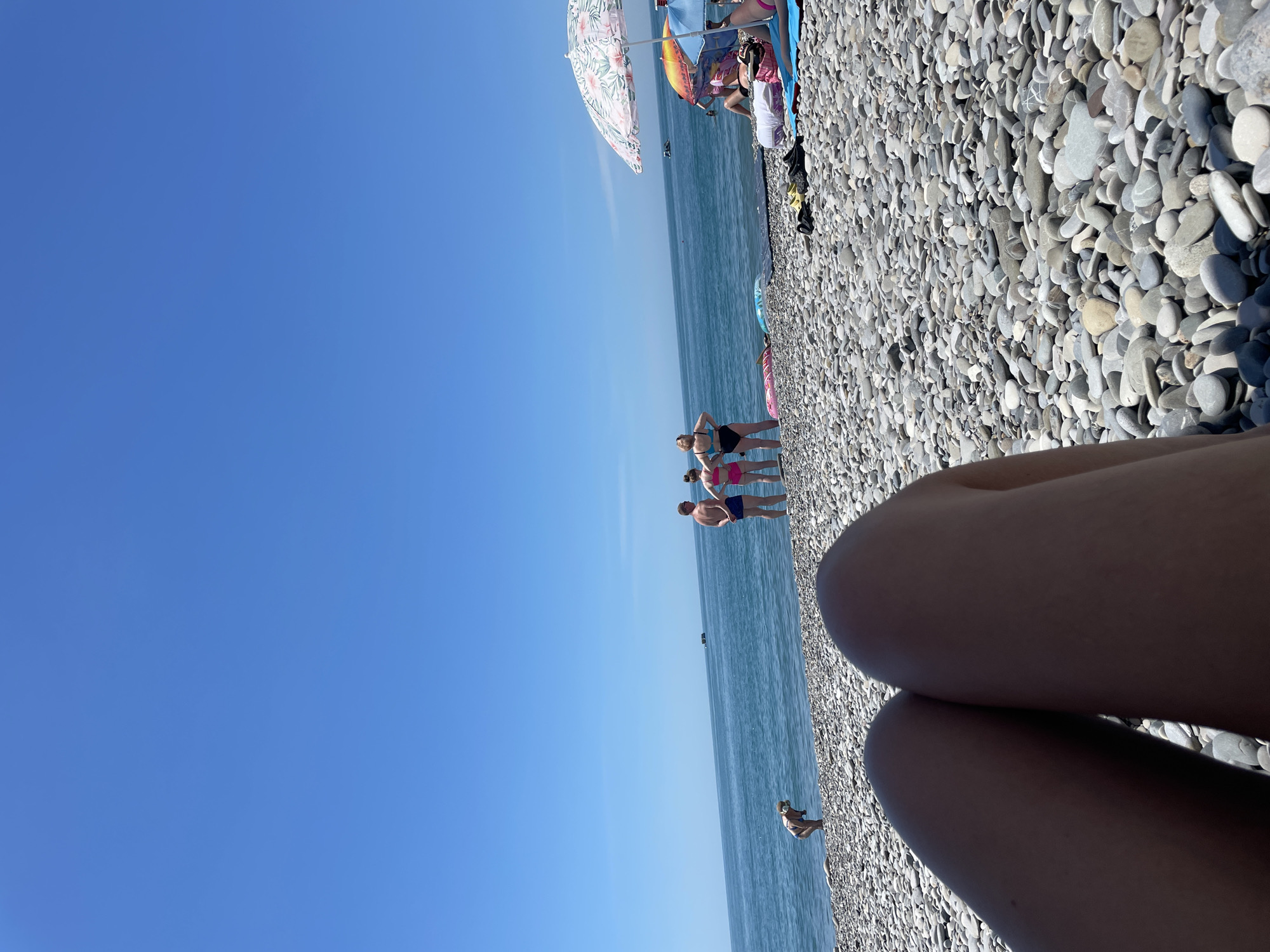 A person's legs are in the foreground of the image, suggesting the photo is taken from a seated or lying position on a beach. The beach is covered in gray pebbles. In the middle ground, three people are standing on the pebble beach near the water's edge, looking out at the sea. One person is bending over near the water. Further into the distance, several beach umbrellas and people are visible, along with a boat on the horizon. The sky is clear and blue. The location is a beach in Unknown, Unknown.