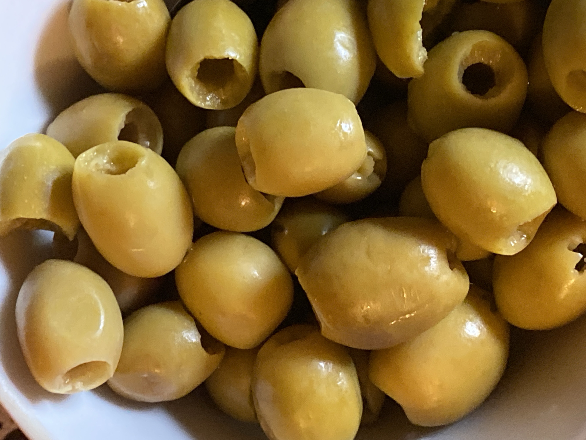 The media file contains a close-up, overhead shot of a bowl filled with pimento-stuffed green olives. The olives are the sole subject, presented in a light-colored bowl, though only a small portion of the bowl is visible at the bottom edge of the frame. The olives are plump and glistening, with a smooth, waxy texture. Each olive has a distinct opening where the pimento has been inserted. The lighting is bright, creating highlights on the shiny surfaces of the olives and casting soft shadows between them. The overall impression is one of a food preparation or presentation. There is no visible text. The context of Voeikovo, Russia, does not provide any direct visual cues within the image itself.