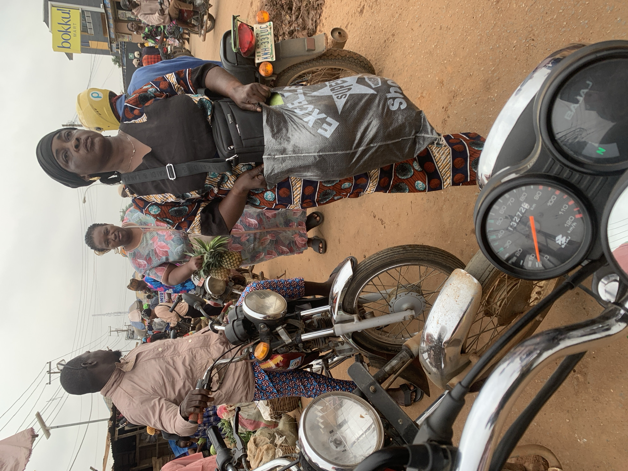 The image captures a street scene in Shagamu, Nigeria, likely a market area given the presence of motorcycles, vendors, and customers. In the foreground, the handlebars and speedometer of a motorcycle are visible, indicating a first-person perspective from someone on a motorcycle.
Several people are present in the scene. A man in a light-colored jacket stands next to a motorcycle, his left hand on the handlebar. To his right, a woman wearing a black headscarf and a vibrantly patterned dress is holding a large black plastic bag with white lettering, "EXCELLENCE SUPER SHOP," in her hands. She also has a black shoulder bag. Behind her, another woman in a patterned dress holds a pineapple. Further in the background, more people are visible, some near other motorcycles and market stalls.
The environment appears to be an unpaved or dusty street. In the background, a yellow sign for "bokku! MART" is visible on a building, suggesting a commercial area. Power lines and poles are present in the sky, typical of an urban or semi-urban setting. The overall atmosphere suggests a busy, bustling day in a Nigerian city.