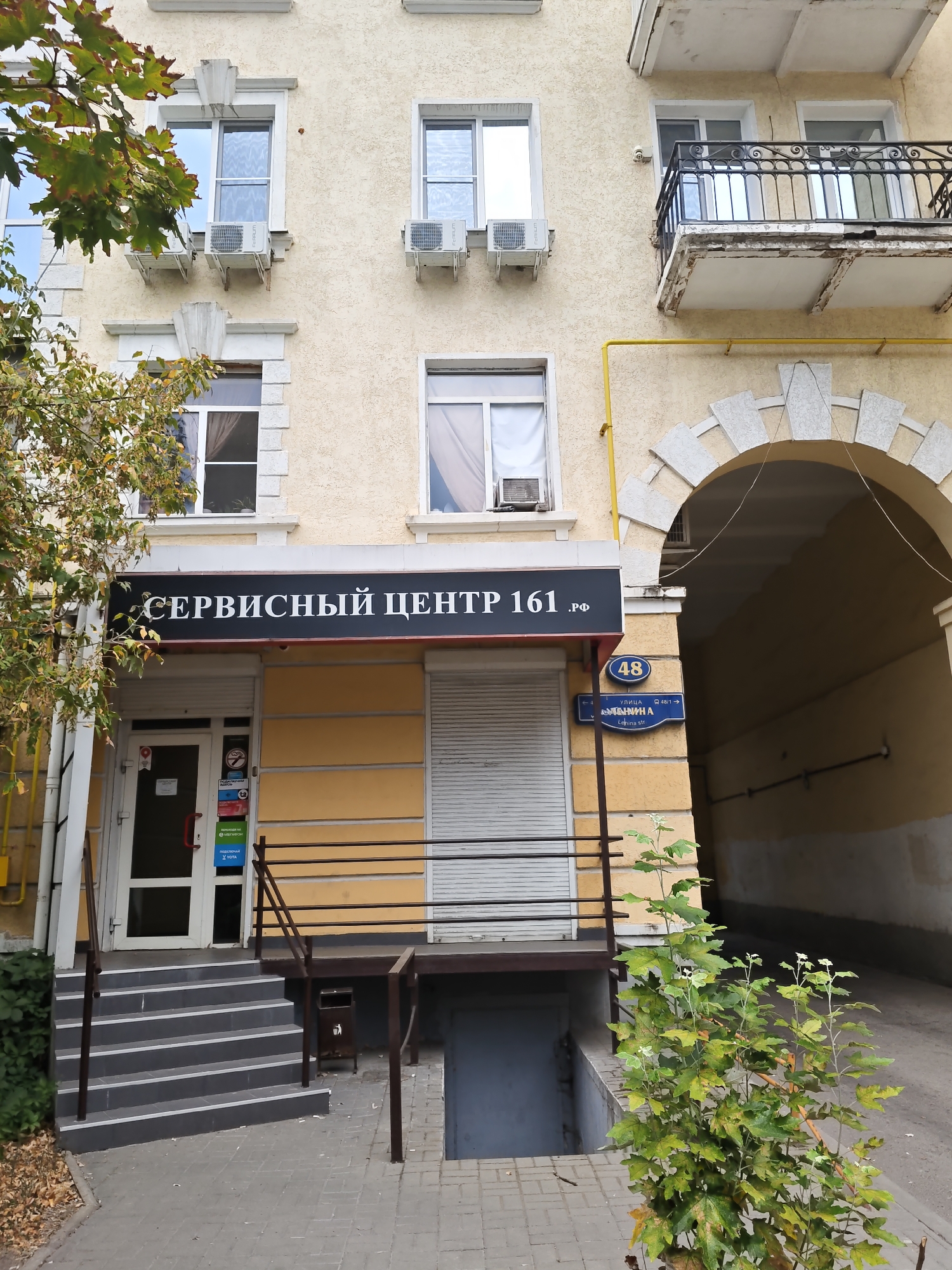 A storefront named "СЕРВИСНЫЙ ЦЕНТР 161 .РФ" (Service Center 161 .RF) is visible on the ground floor of a building. The storefront has a glass door and is protected by a rolling metal shutter. To the left of the entrance, there are stairs leading up to the door, and a green bush is planted to the right. Above the storefront, several windows with air conditioning units are visible on the building's facade. A street sign with the number "48" and Cyrillic text indicates the location is at "улица Комарова" (Komarova Street), a street in Rostov-on-Don, Russia. The building appears to be old, with some signs of wear and tear on the balcony railings. A yellow gas pipe runs vertically up the side of the building.