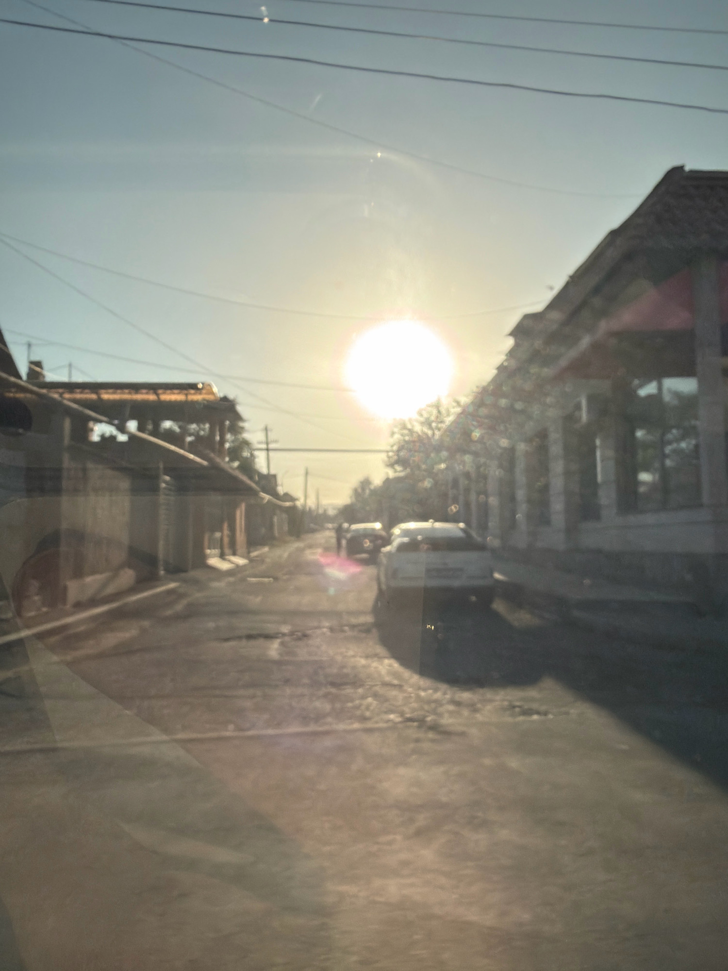 A sun-drenched, slightly blurry street scene in Yerevan, Armenia, depicts a road with buildings on either side. The sun is high in the sky, creating lens flare and glare on what appears to be a car windshield, suggesting the photo was taken from inside a vehicle. Two cars are visible on the road, one white and one darker, both appearing to be in motion or parked. The buildings have a weathered appearance, with awnings and traditional roofing. Power lines are strung across the sky. The road surface is uneven and appears to be paved. The overall impression is a candid snapshot of everyday life on a sunny day in the city.