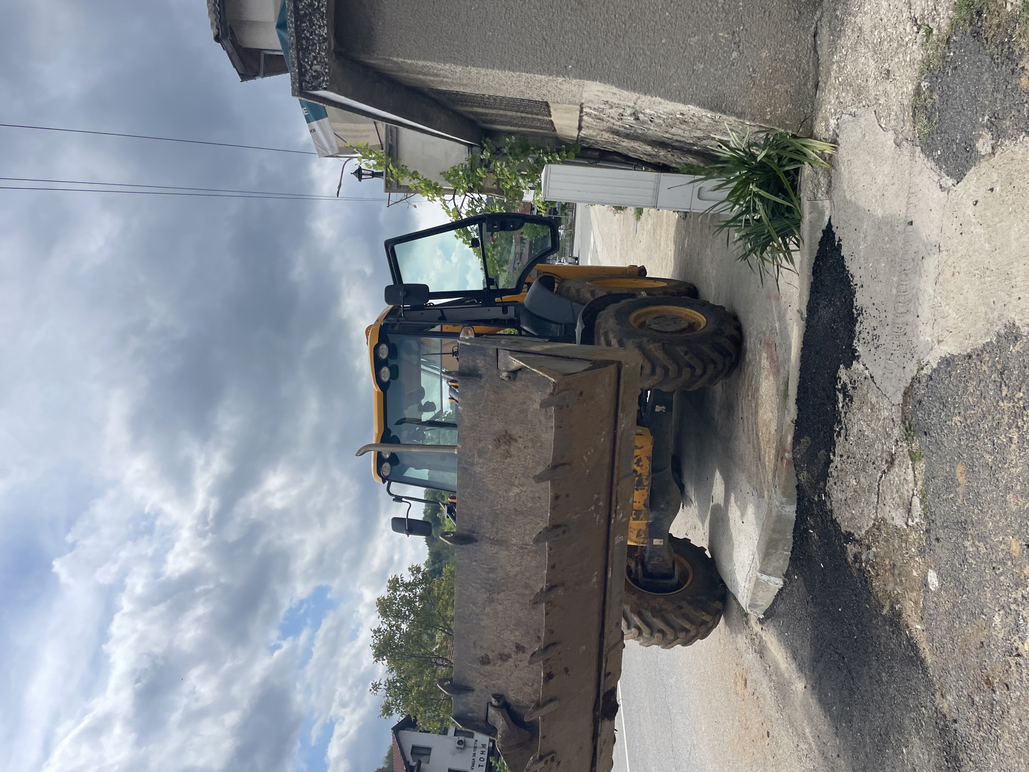 A yellow backhoe is parked on a narrow street in Zlatograd, Bulgaria. The backhoe's front bucket is extended and facing the viewer. The vehicle is positioned against a building wall with overgrown foliage. The street surface shows evidence of recent asphalt work. The sky is overcast with heavy clouds.