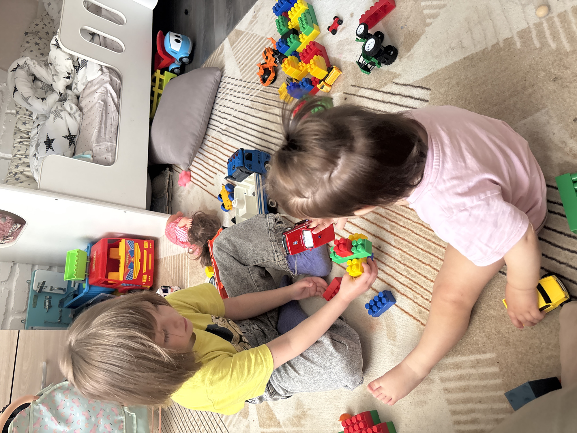 Two children are playing with toys on a rug in a room. One child wears a yellow shirt and grey jeans. The other child wears a pink shirt. There are various toys scattered around them, including building blocks and toy vehicles. A bed and other furniture are visible in the background. The image appears to be taken indoors. The location is Chaikovsky, Russia.