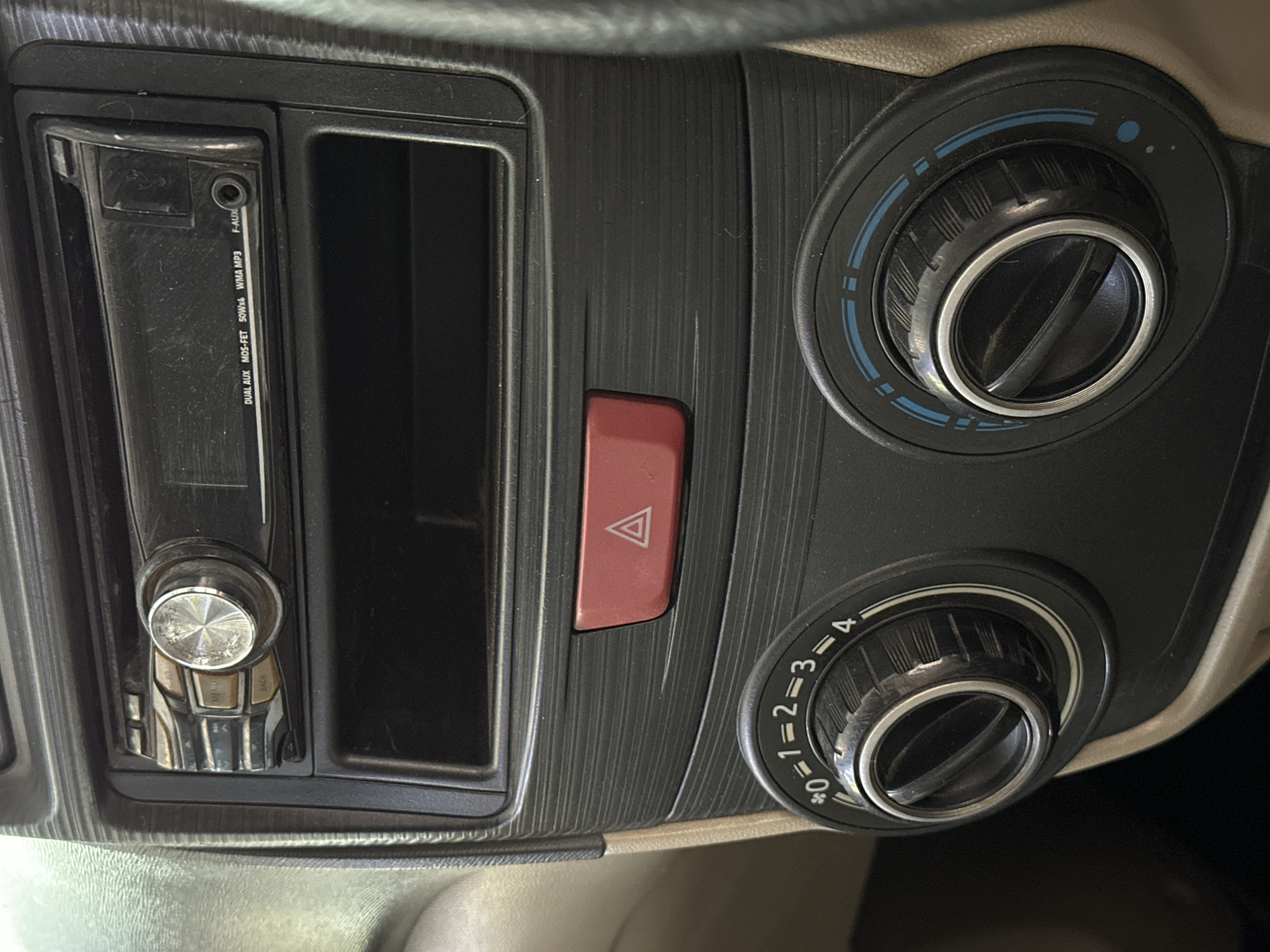 The image shows a close-up view of the dashboard of a car, specifically the center console area. Prominently featured is a car stereo system with buttons and a display, below which is an empty storage slot. To the left of the storage slot, there are two rotary knobs with markings. The left knob has numerical markings from 0 to 4, with a snowflake symbol next to the 0, suggesting fan speed control. The right knob has blue and light blue indicators around its circumference, possibly for temperature or air direction. In the middle of the console, between the stereo and the knobs, is a red hazard light button with a white triangle symbol. The dashboard material appears to be dark plastic with a textured, horizontally ribbed finish. The surrounding environment is out of focus but suggests the interior of a vehicle. The specific location in Bulakrejo, Indonesia, is not visually evident in the provided image.