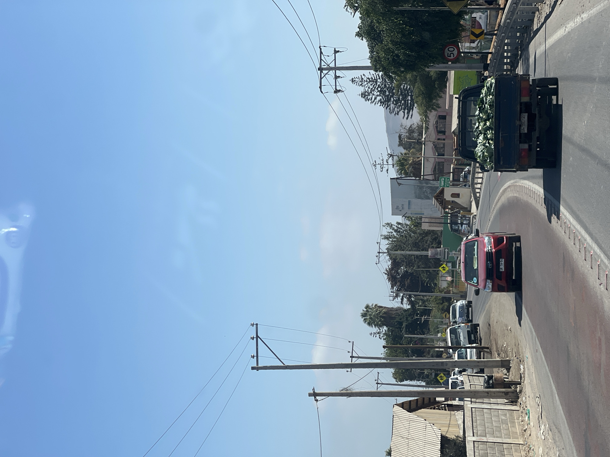 The image shows a street scene in Arica, Chile, on a sunny day. The view is from the roadside, looking down the road, with a clear blue sky above. A red car is driving up the street, which has a line of parked white cars along the side. Further up the street is a dark pickup truck filled with what appears to be green produce. There are power poles along the side of the road and buildings and trees visible in the background. Speed limit sign is visible.
