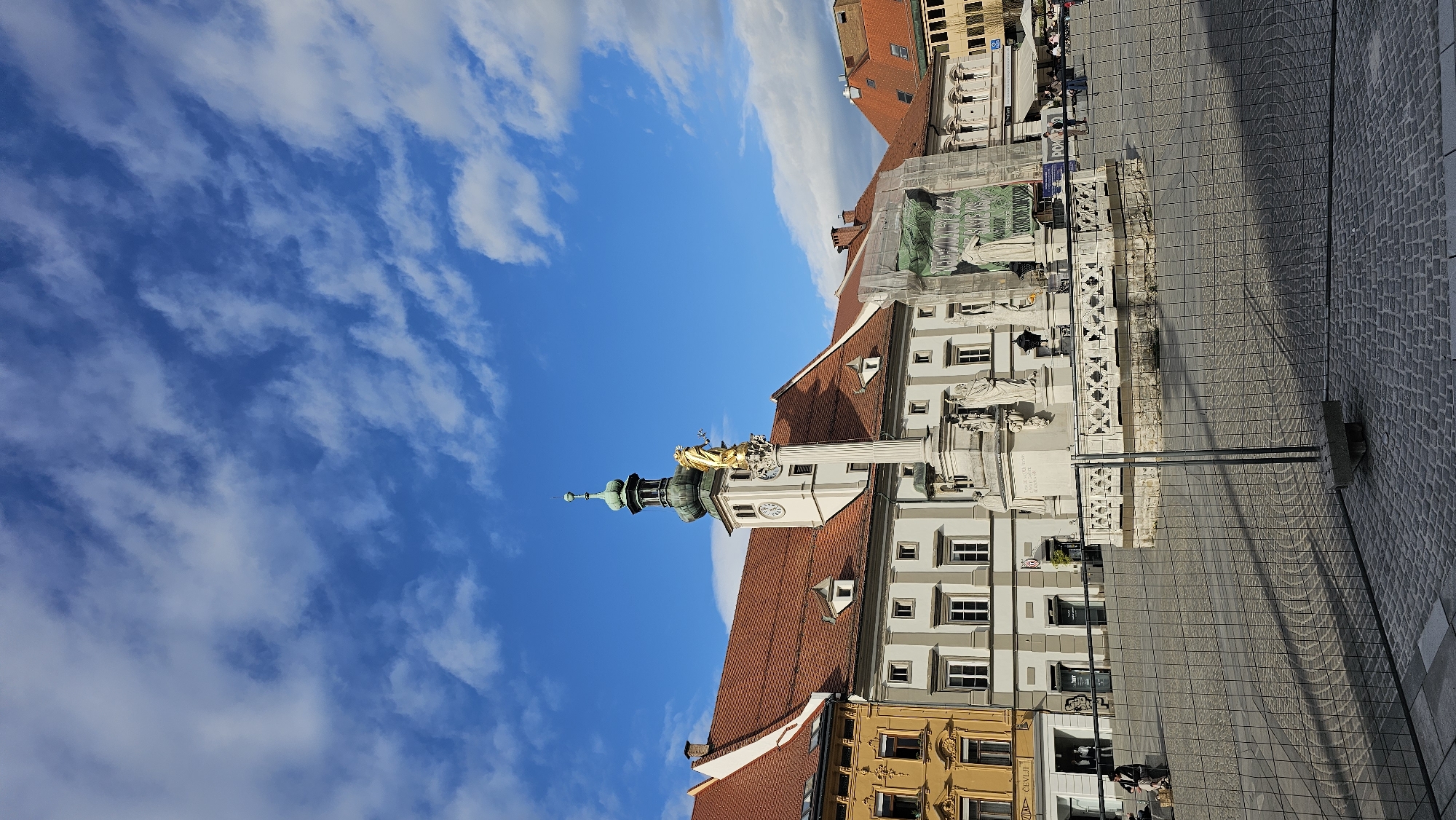 The image depicts the Main Square (Glavni trg) in Maribor, Slovenia, featuring the Plague Column, also known as the Monument to the Holy Virgin. The column is a prominent white structure with a golden statue on top, likely representing the Virgin Mary. Buildings with orange tile roofs and light-colored facades frame the square. A paved pedestrian area stretches out in front of the monument. The sky above is a bright blue with scattered white clouds. People are visible walking in the background.