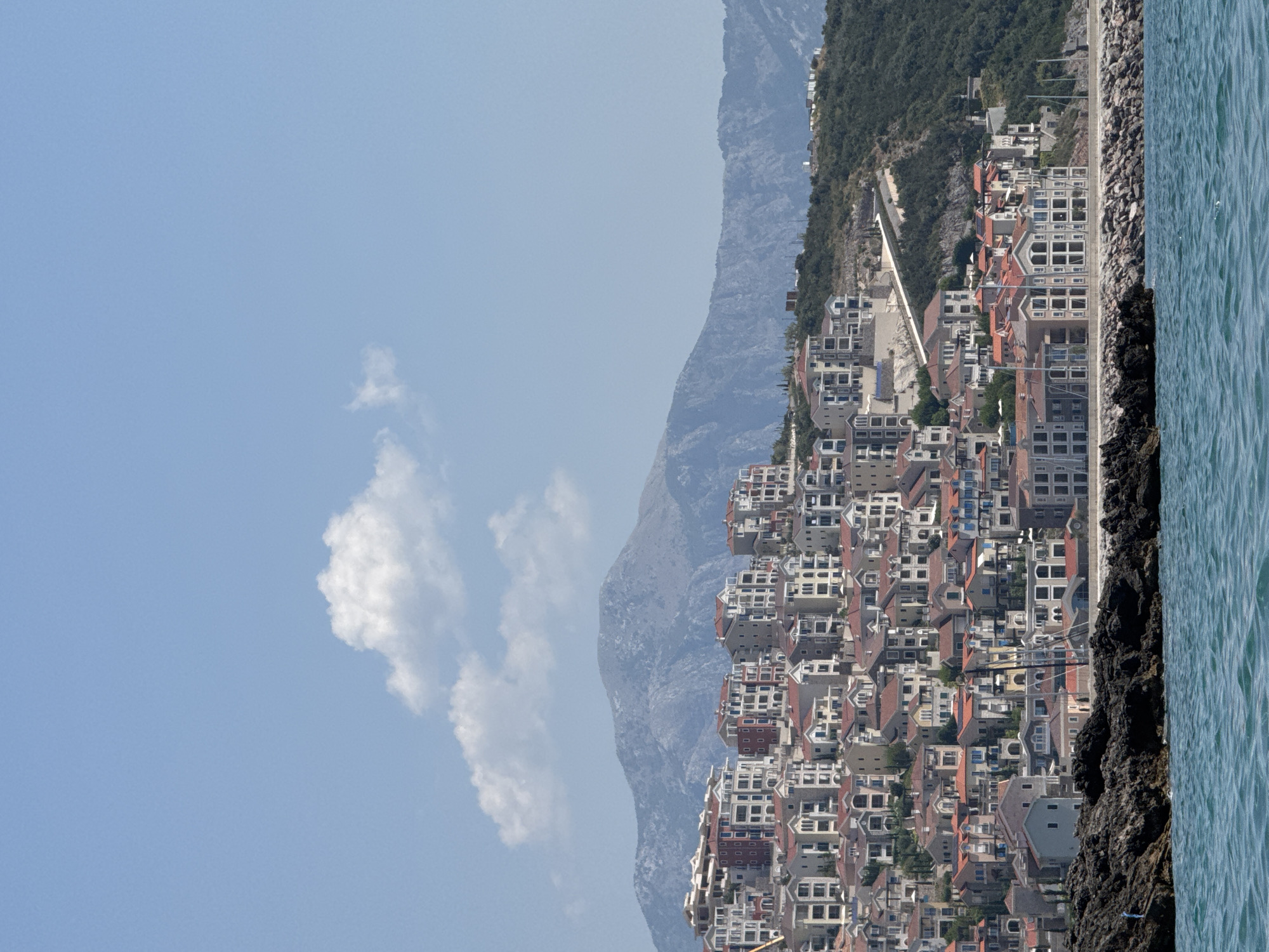 The image shows a dense cluster of multi-story buildings with red-tiled roofs situated on a hillside overlooking a body of blue water. The architecture appears modern, with many balconies and large windows. In the foreground, dark, rough rocks line the edge of the water, forming a natural barrier. Behind the buildings, a winding road is visible ascending the hill, and further in the background, a large, rugged mountain range dominates the horizon under a clear blue sky with a few wispy clouds. The scene depicts a coastal town, likely in a Mediterranean climate, possibly Radovići, Montenegro, given the architectural style and mountainous backdrop often associated with the region. There are no people visible in the image. The overall impression is one of a developed residential area integrated into a natural, hilly landscape.