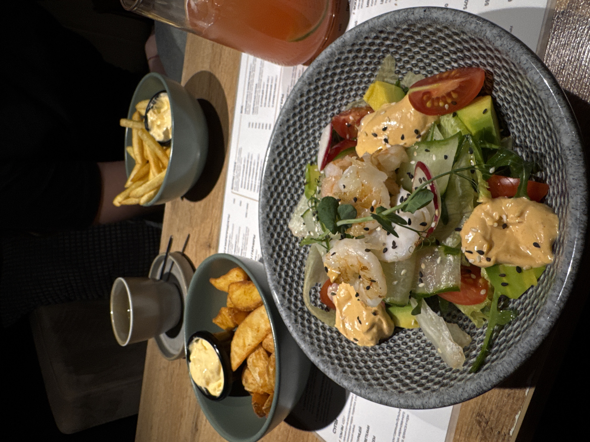 The image shows a dining table in Chaikovsky, Russia. A salad with shrimp, tomatoes, cucumbers, avocado, and a creamy dressing is in a gray patterned bowl. There are two smaller bowls, each containing french fries and a sauce. A cup is visible next to one of the french fry bowls. A drink in a glass pitcher is placed near the salad bowl. A person wearing black clothing is seated at the table, and a menu is visible on the table. The table is wooden.