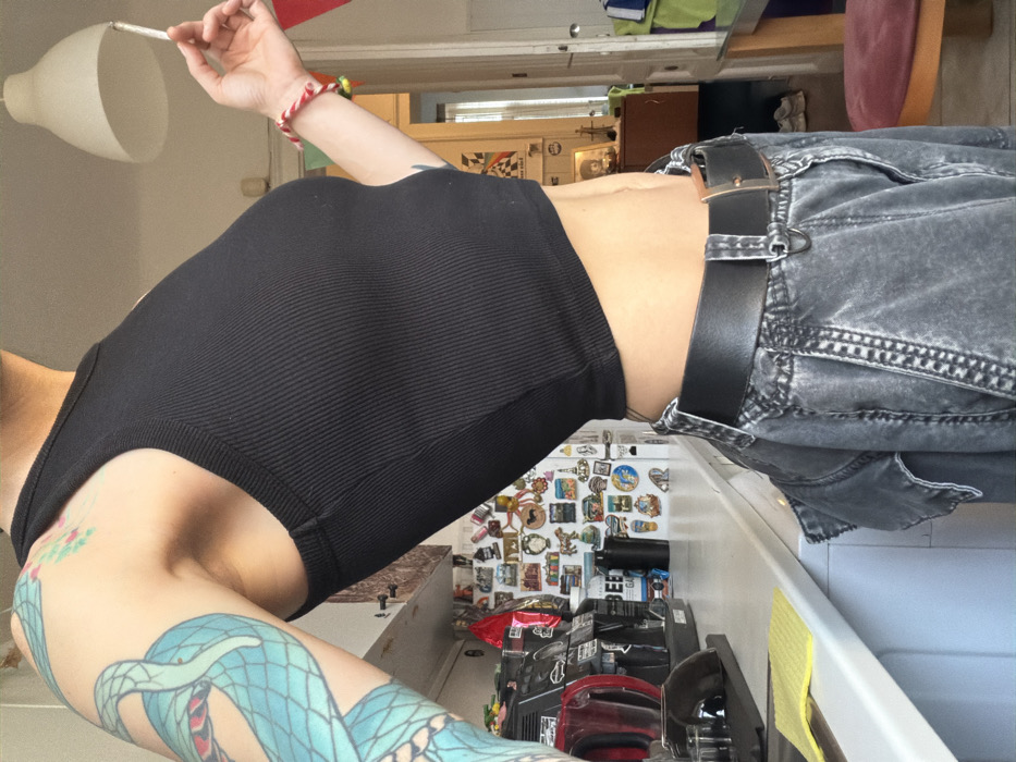 An individual is depicted from the mid-chest to the upper thighs, standing in what appears to be a residential kitchen or dining area in Sofia, Bulgaria. The person wears a black ribbed sleeveless crop top, exposing the midriff, and faded grey denim cargo pants with a black leather belt and a copper-toned buckle. The visible left arm is extensively tattooed with a large, light blue serpentine design and smaller green and pink floral motifs. The right hand holds a lit cigarette between the index and middle fingers. On the right wrist, a red and white patterned bracelet and a multicolored beaded bracelet are visible.
The background to the left features a white kitchen counter with a yellow sponge and a black and red appliance, possibly a coffee maker. Behind the counter, a white refrigerator is densely covered with various souvenir magnets. To the right, a doorway opens to another room, where a wooden table and a red-cushioned stool are partially visible. A white, inverted cone-shaped pendant light fixture hangs from the ceiling.