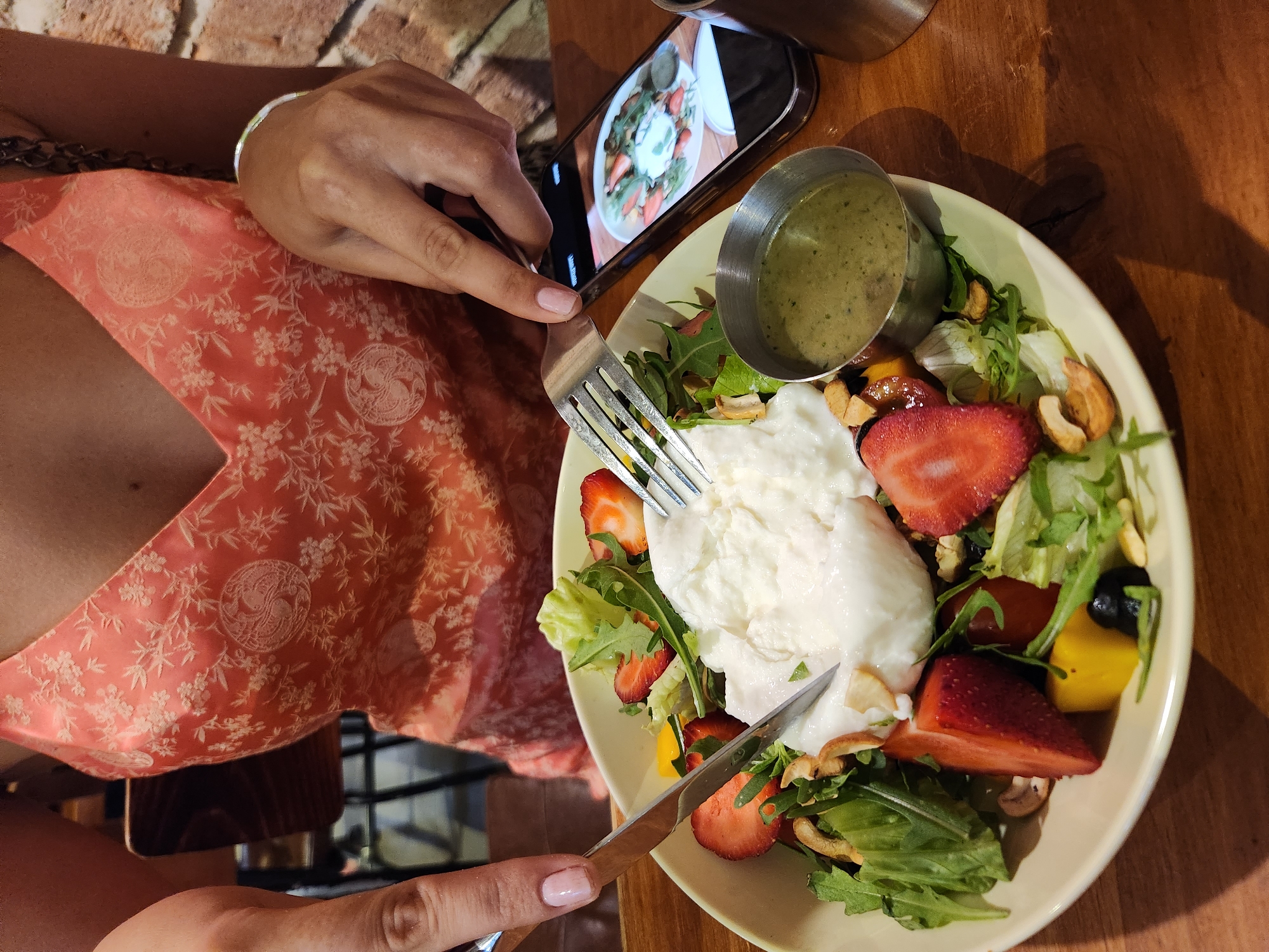 A close-up, overhead shot shows a person's hands cutting into a burrata salad in a restaurant in Da Nang, Vietnam. The salad, served in a light yellow bowl, contains burrata cheese, strawberries, arugula, lettuce, cashews, yellow bell peppers, and olives. A small metal cup of salad dressing sits to the right of the bowl. A smartphone displaying an image of the salad is placed to the right of the bowl. The person is wearing a coral-colored dress with a floral pattern. The background shows a brick wall and a wooden table.
