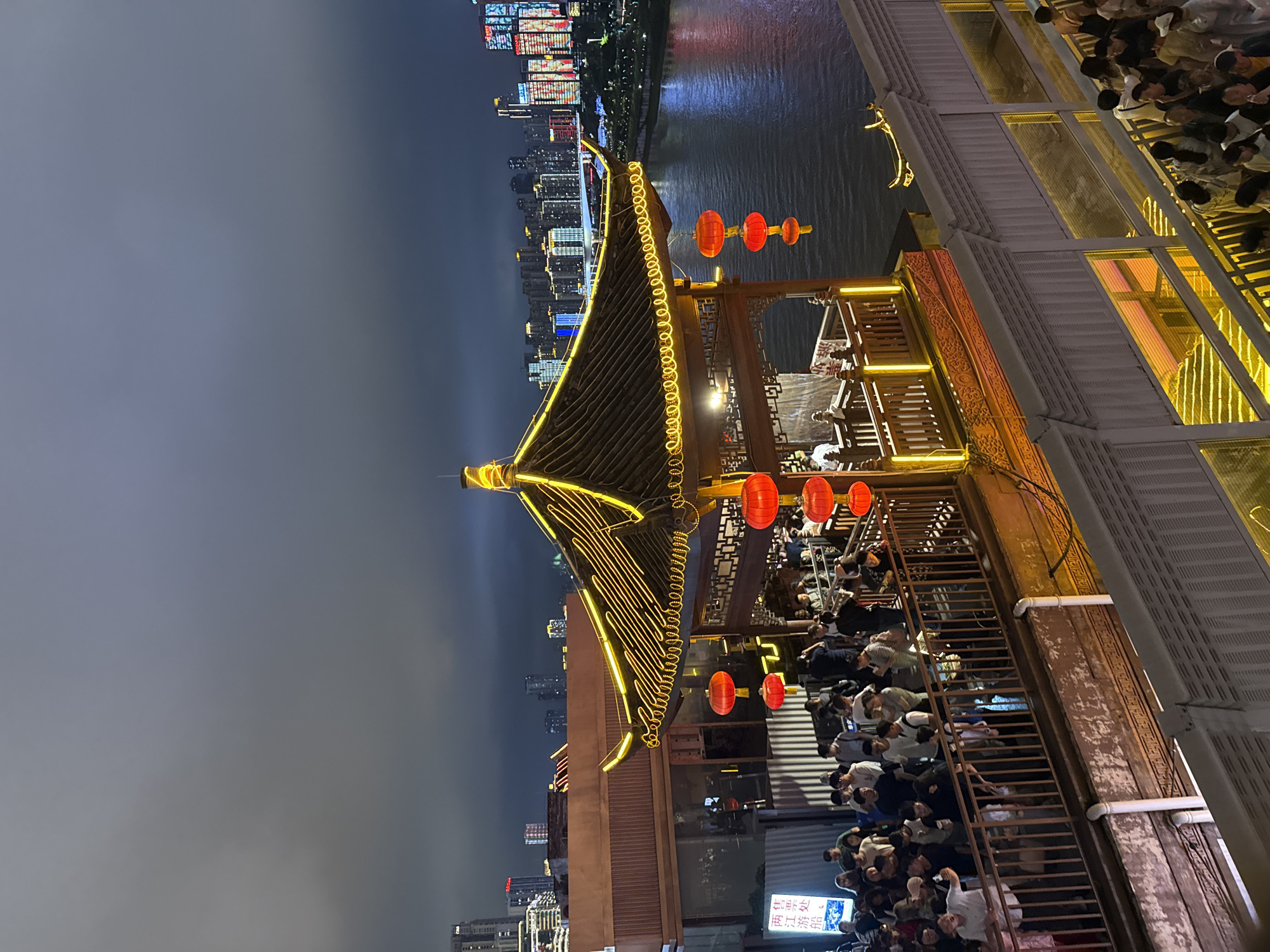 This media file captures a nighttime scene in Yuzhong District, China, likely during an evening gathering or event. The focal point is a multi-tiered structure with traditional Chinese architectural elements, including a dark, sloped roof adorned with glowing yellow neon lights. Red lanterns are strategically placed around the structure, adding to the festive ambiance.
A river flows in the background, reflecting the vibrant lights of a city skyline that stretches across the horizon. The buildings in the distance are illuminated with a colorful array of lights, suggesting a bustling urban environment.
The structure itself appears to be a restaurant or viewing platform, as indicated by the presence of numerous people gathered on its various levels. They are seated at tables, standing and socializing, and some are looking out towards the river and city. The atmosphere seems lively and convivial, with many people appearing engaged in conversation and enjoying the night.
The time of day is clearly night, with the sky a dark, muted gray, possibly indicating overcast conditions or a hazy atmosphere. The lighting from the city and the structure itself dominates the scene, casting warm glows and sharp shadows.
Visible text can be seen on a sign at the bottom right of the frame, featuring Chinese characters. The characters appear to be part of a business name or advertisement, contributing to the sense of place within China. The overall impression is one of a vibrant, culturally rich urban setting at night, with a strong emphasis on communal enjoyment and a blend of traditional and modern aesthetics.