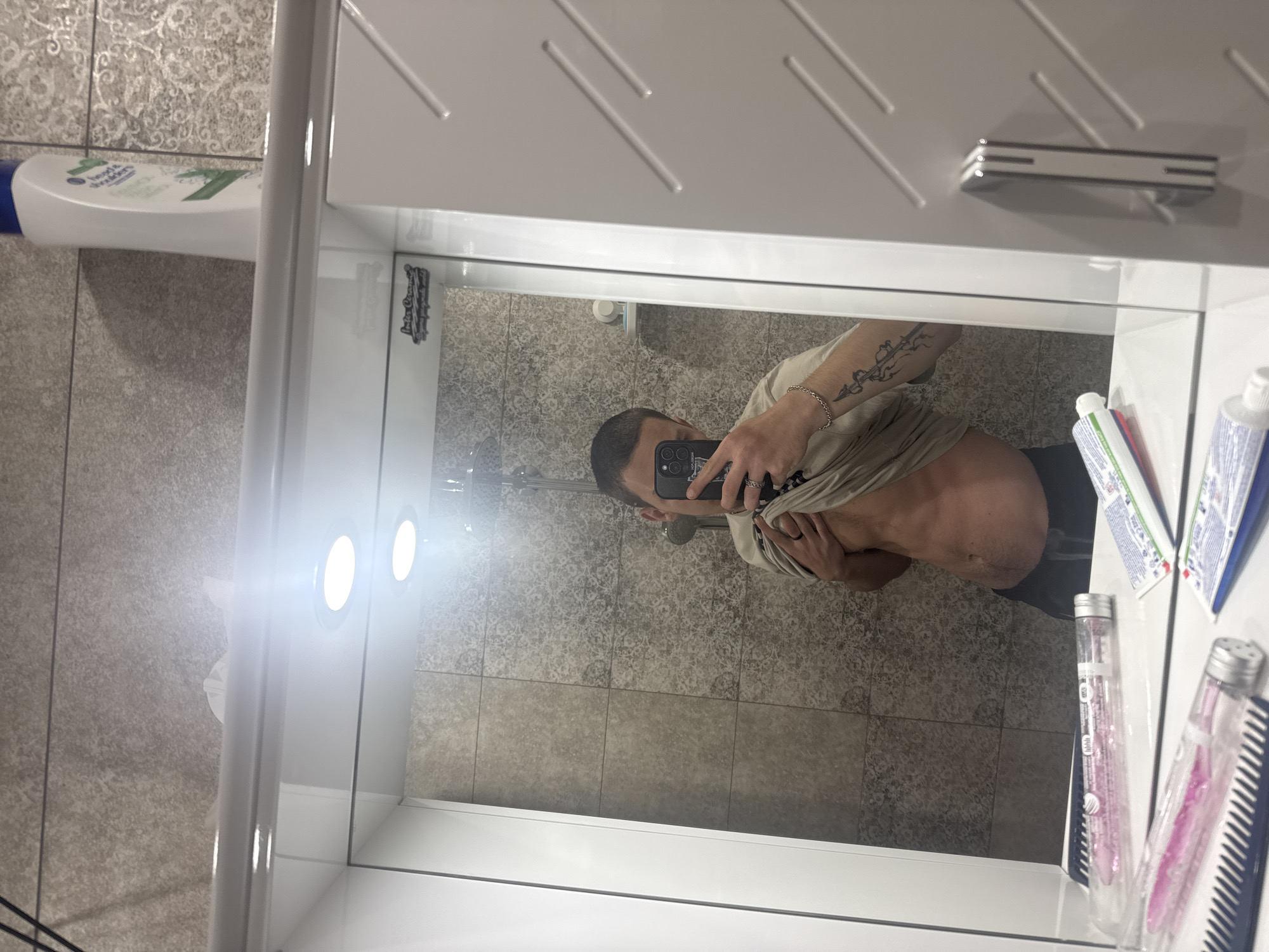 The image displays a mirror selfie taken by a person in a bathroom, presented with an unusual vertical orientation. The scene is dominated by a large mirror reflecting a portion of the room and the individual.
The primary subject is a male, visible from the chest down, holding a black smartphone with his right hand to capture the reflection. He has short hair and is wearing a light-colored t-shirt that is lifted to reveal his bare torso and lower abdomen. A silver chain bracelet adorns his left wrist, and a distinctive tattoo of swirling, organic patterns is visible on his left forearm. A ring is also visible on his right hand.
The bathroom features tiled walls; the upper section has a decorative, patterned grey tile, while the lower section consists of plain, larger rectangular grey tiles. A white cabinet or shelving unit, likely above a sink (not fully visible), is prominent. Integrated into this unit are two bright, circular light fixtures, creating significant glare and reflections on the mirror's surface.
Various personal items are scattered around the bathroom. To the left, on a shelf or ledge, rests a white bottle with a blue cap, clearly identifiable as "Head & Shoulders" shampoo or conditioner. On the right side, inside the cabinet's reflection, are several tubes, including toothpaste, and two clear plastic containers holding combs. A small black sticker reflecting in the mirror reads "Los Angeles California" along with other less legible text.
The activity depicted is the act of taking a mirror selfie, possibly to document fitness progress or simply a casual moment. The strong artificial lighting suggests an indoor setting, and the time of day is not explicitly evident.