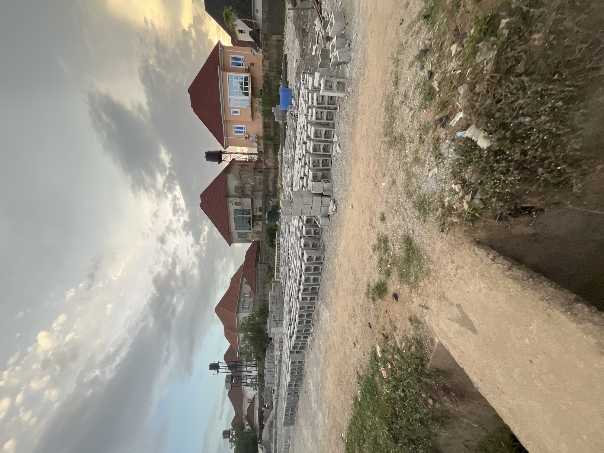 The image displays a developing residential area in Abuja, Nigeria. The foreground features an unpaved dirt ground, transitioning into patches of green vegetation and some visible litter on the right. A concrete curb is present at the bottom right.
In the midground, large quantities of concrete blocks are neatly stacked in rows, indicating ongoing construction or a building material depot. Beyond these stacks, several residential houses with reddish-orange pitched roofs and light-colored facades are visible. Some houses feature blue-tinted windows, and one has a black water tank elevated on a metal stand next to it. Other buildings are visible further in the background, partially obscured by trees.
The upper portion of the image is dominated by the sky, filled with a mix of light and dark clouds. Golden light filters through the clouds, particularly on the left, suggesting either sunrise or sunset. The overall atmosphere is quiet, with no people or specific activities evident. There is no visible text in the image.