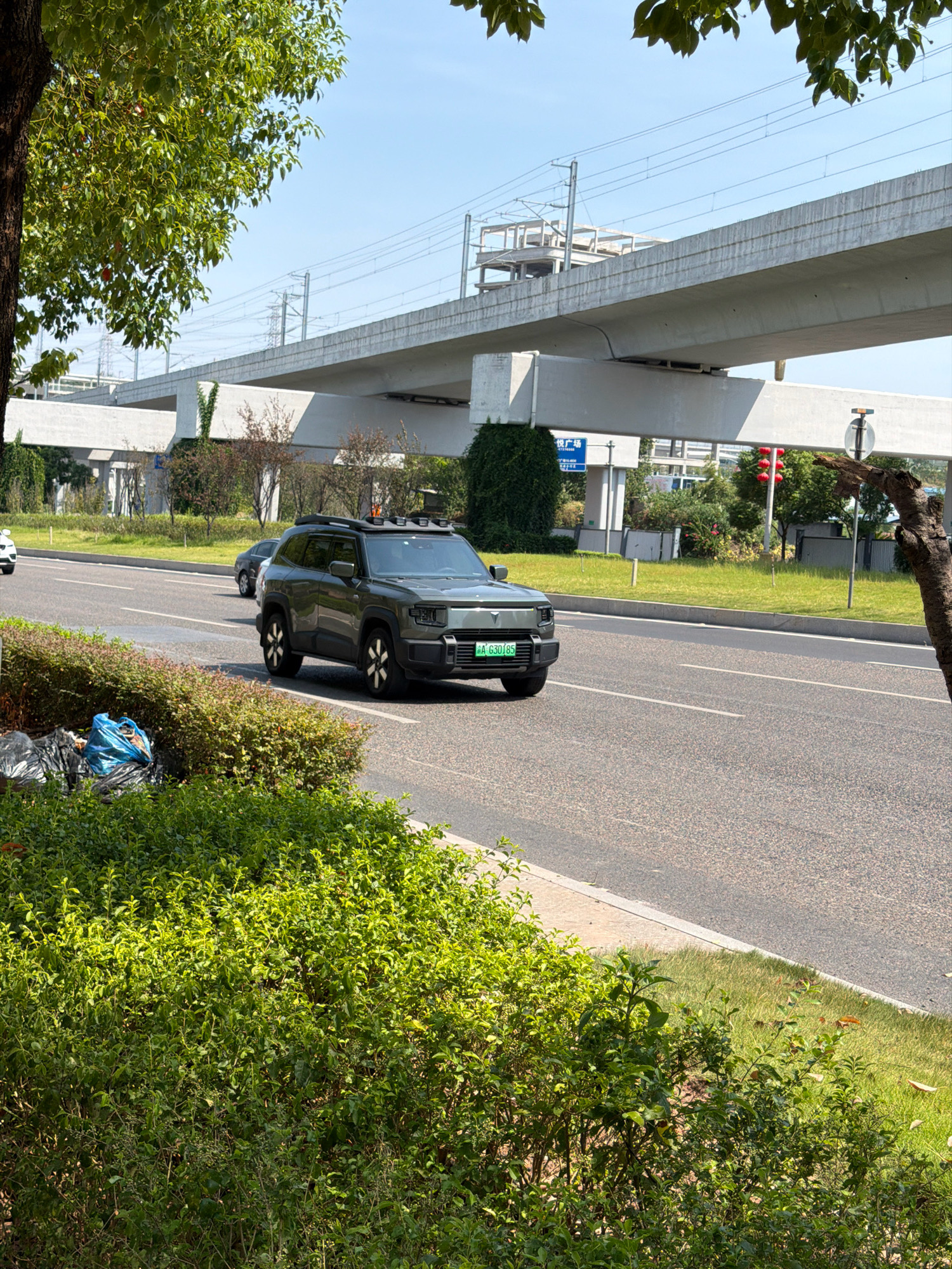 A dark green SUV with a license plate that reads "A 630185" is driving on a paved road in Jiangjin, China. The SUV has a roof rack with several lights. The road is lined with green bushes and grass. In the background, a concrete overpass stretches across the scene. The sky is blue and clear. To the left of the road, there are several black trash bags piled up next to a tree. A white car is visible in the distance on the left side of the road. The image appears to have been taken during daylight.