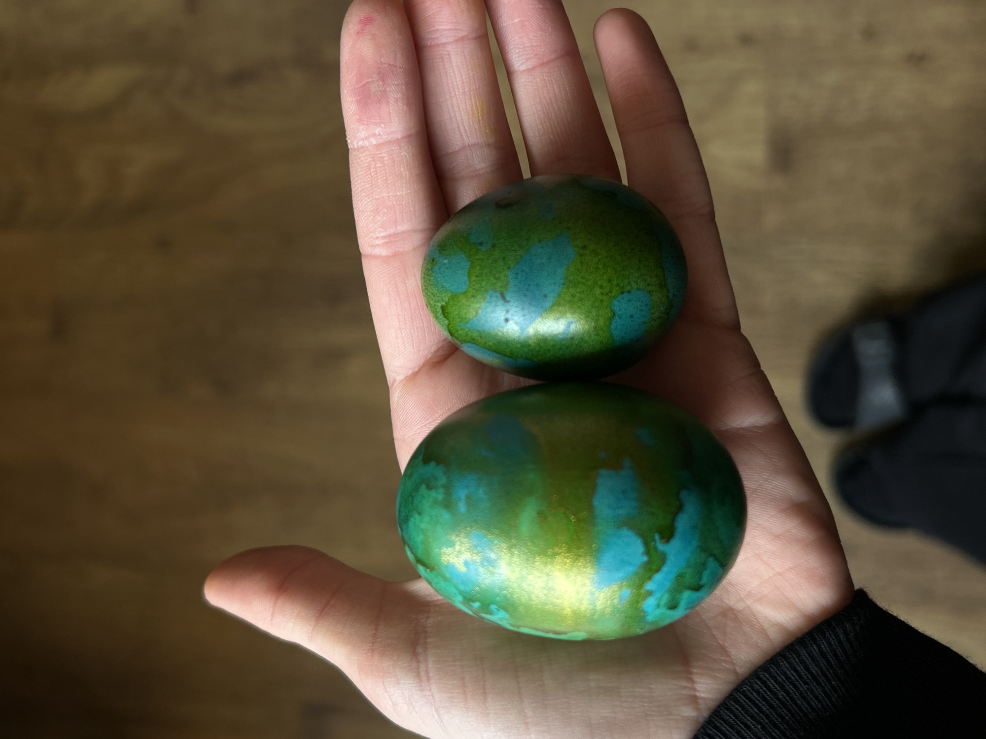 The image shows a person's open palm holding two green and blue colored, egg-shaped objects. The objects have a mottled appearance. The background is out of focus, but appears to be a wooden surface. Part of a black garment is visible in the lower right corner, and out-of-focus footwear is visible at the top right corner. There is no evidence to suggest a location in Krumovo, Bulgaria.