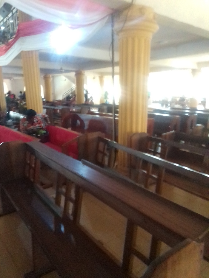 The image shows the interior of a building in Auchi, Nigeria, located at latitude 7.066 and longitude 6.257. The image is taken from the perspective of someone standing in the pews of a church, looking towards the front of the building. The pews are made of dark wood and have red cushioned seats. There are several large pillars supporting the roof of the building, and the walls are painted a light beige color. The image appears to have been taken during a service, as there are people sitting in the pews, but they are out of focus. A white and red banner hangs above the entrance to the building. A row of windows is visible on the opposite side of the room, and natural light shines in through the windows.