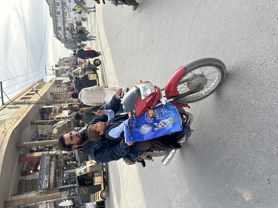 An outdoor, eye-level photograph depicts a man and a child riding a red motorcycle on an asphalt road in Quetta City Tehsil, Pakistan, under an overcast sky. A man with a dark beard, wearing a dark blue jacket and dark pants, operates the red Honda "Sports" motorcycle. Seated in front of him is a young male child, dressed in a light blue long-sleeved shirt and a dark vest, with his right index finger pointed upwards and his mouth agape. A blue plastic shopping bag, branded "Mihan Milk," hangs from the motorcycle's handlebars. The street features multiple pedestrians and vehicles, including several beige and black auto-rickshaws and other motorcycles. Multi-story commercial buildings line both sides of the street, displaying various storefronts with Urdu signage, some indicating "Catering" and "Clinic." A dense network of overhead electrical wires is visible against the light gray sky. A person wearing a yellow safety vest is discernible in the distant right background among other pedestrians.
