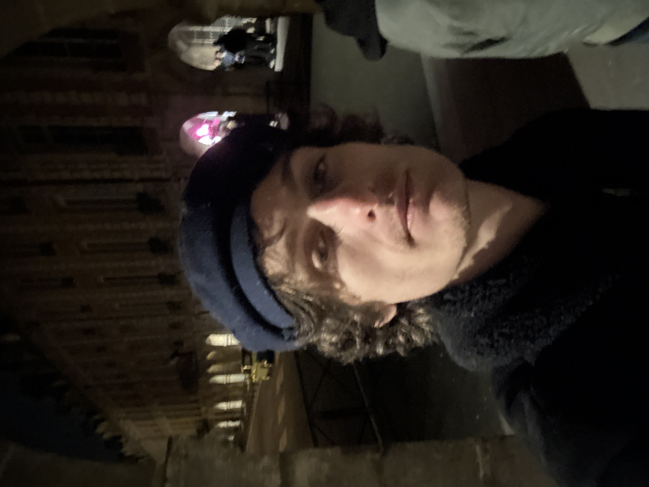 A person wearing a dark cap and jacket takes a selfie. The face is lit from the front, showing stubble, curly hair and a nose towards the camera. Behind, a large architectural stone structure rises into the dark. Arched openings and decorated walls suggest an old building. A colorful glow is visible through one arch, while figures stand silhouetted in another.