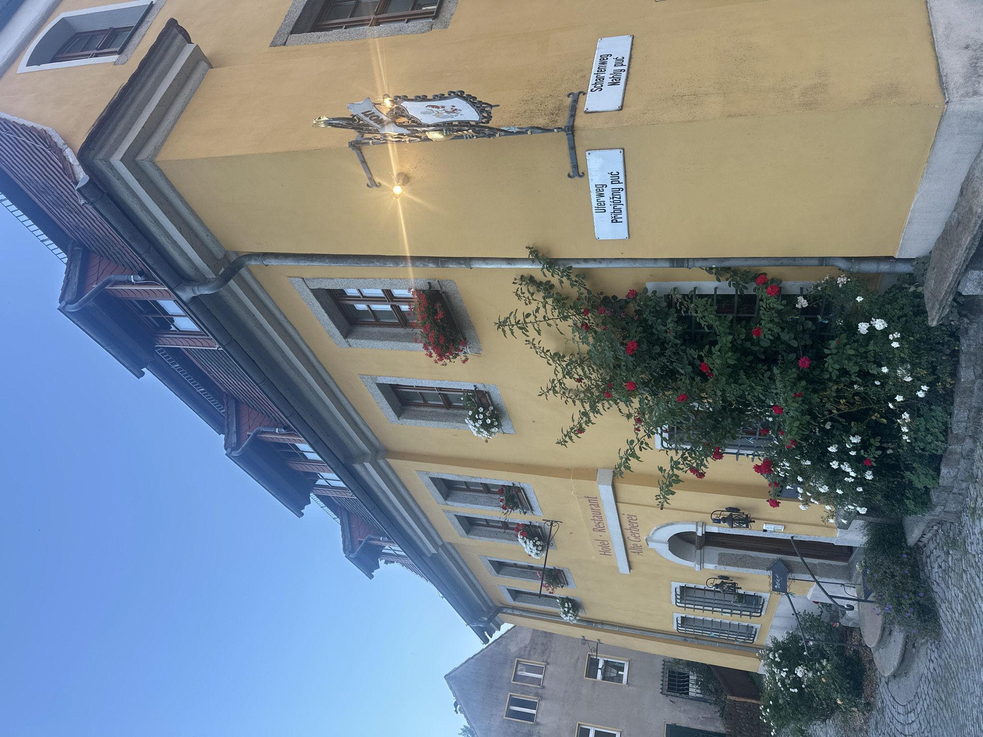 This image captures a corner view of a yellow building in Bautzen, Germany, identified by street signs as "Uferweg" (with Sorbian text "PřiBrjoźny puć") and "Scharfenweg" (with Sorbian text "Nahły puć"). The building appears to be a hotel and restaurant named "Alte Gerberei." It features multiple windows with flower boxes containing red and white flowers. A decorative sign with a crest hangs from an ornate street lamp. The building's architecture includes a tiled roof and stone accents. Lush red and white roses climb the side of the building, and a cobblestone path leads to the entrance. A stone bench sits in front of the building. The sky is a clear, pale blue, suggesting it is either early morning or late evening. The environment is quiet and appears to be a historic part of the city. There are no people visible in the image.
