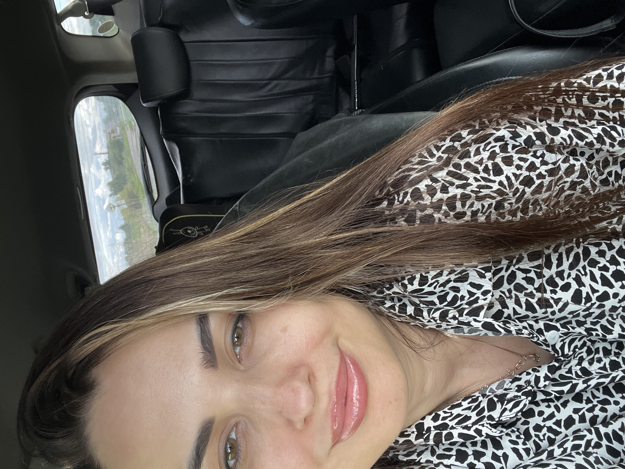 A woman with long, brown hair with light streaks, wearing a black and white patterned shirt and a gold-colored necklace, is depicted from the upper chest upwards, positioned within the left two-thirds of the frame. Her dark brown eyes are open, and her lips, coated with a glossy, light-colored product, are curved in a subtle smile. She is seated inside a vehicle, with the dark interior, including black leather seats and a headrest, visible behind her on the right. A child's car seat with a white bunny illustration is partially visible on the back seat to her left. Through the rear window, a rural landscape is visible under a partly cloudy sky, featuring green trees, a dirt road, and a distant reddish-brown industrial chimney. The location is in the city of Unknown, Unknown.
