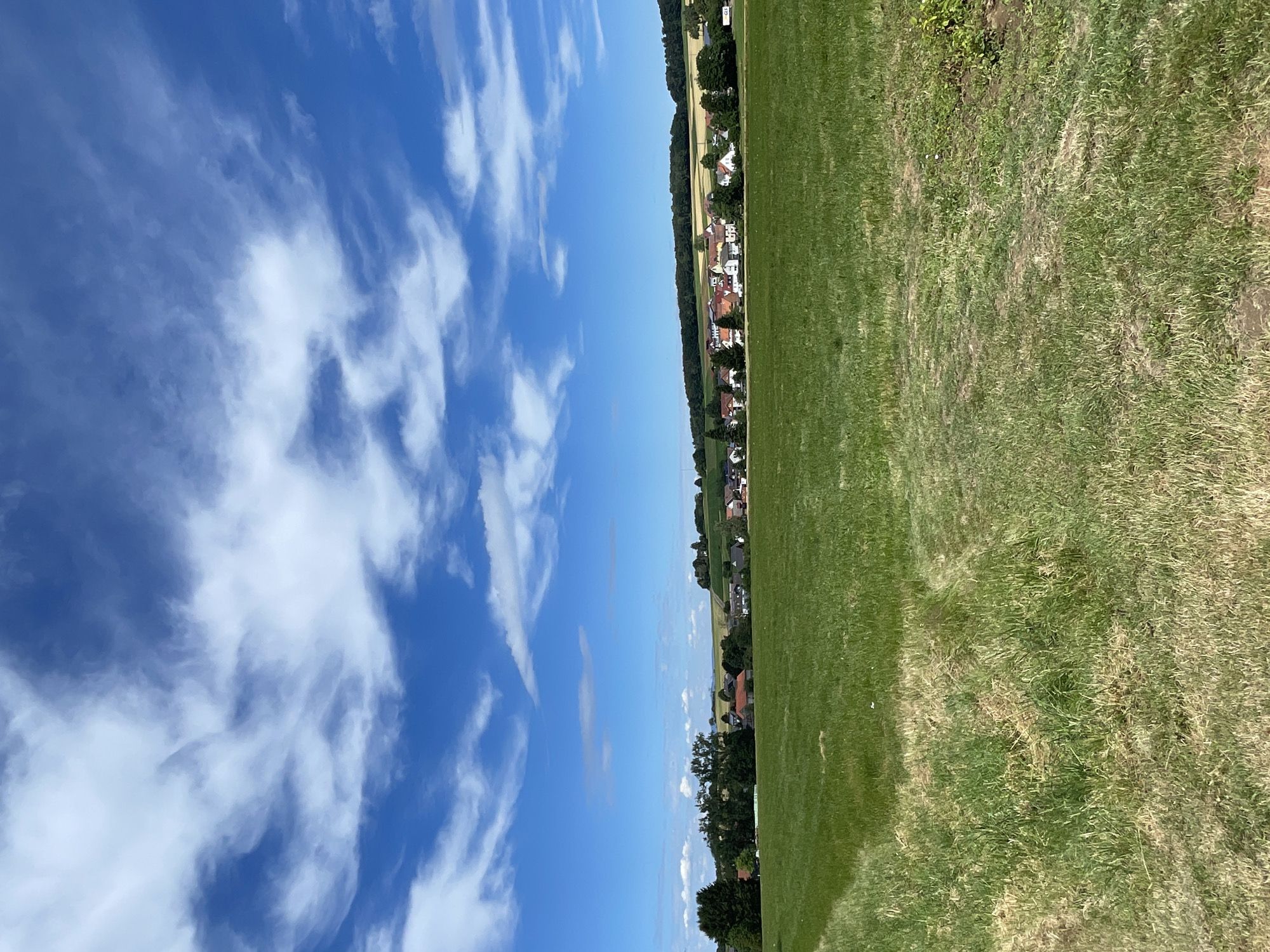 A landscape photograph shows a rural village nestled on a gentle slope under a bright blue sky dotted with white, wispy clouds. In the foreground, a vast, green field dominates the lower half of the image, with slightly drier patches and tire tracks suggesting agricultural use. The middle ground reveals a cluster of houses with red-tiled roofs and white walls, forming a village that extends along the curve of the land. Trees and vegetation surround the village, and a dark green line of forest borders the horizon. To the far left, a large, dense tree stands prominently. The overall scene depicts a peaceful, sunlit day in a German countryside setting, likely Christerode, Germany, given the context. There are no people or animals visible in the frame.