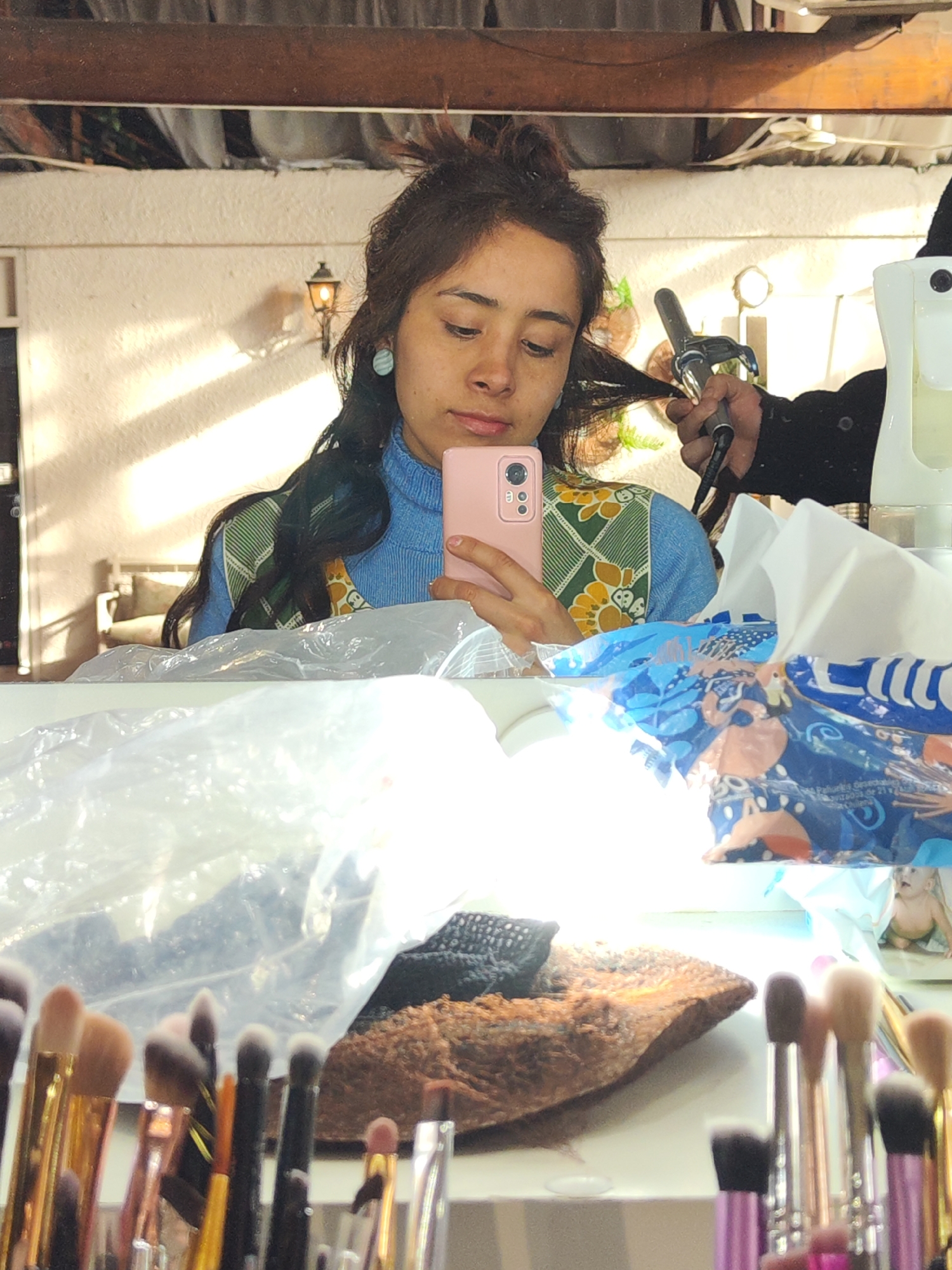 In a bathroom or vanity area in Santiago, Chile, a young woman with dark, wavy hair tied up is reflected in a mirror, holding a pink smartphone in her right hand and using a curling iron on a section of her hair with her left hand. She wears a blue turtleneck sweater and a patterned vest with green, yellow, and white designs. Several makeup brushes are visible in the foreground, some in a clear container. A white tissue box with a blue and patterned wrapper sits to her right, and a white spray bottle is also present. In the background, a lamp with an orange glow is mounted on a textured, light-colored wall, with sunlight casting diagonal shadows.