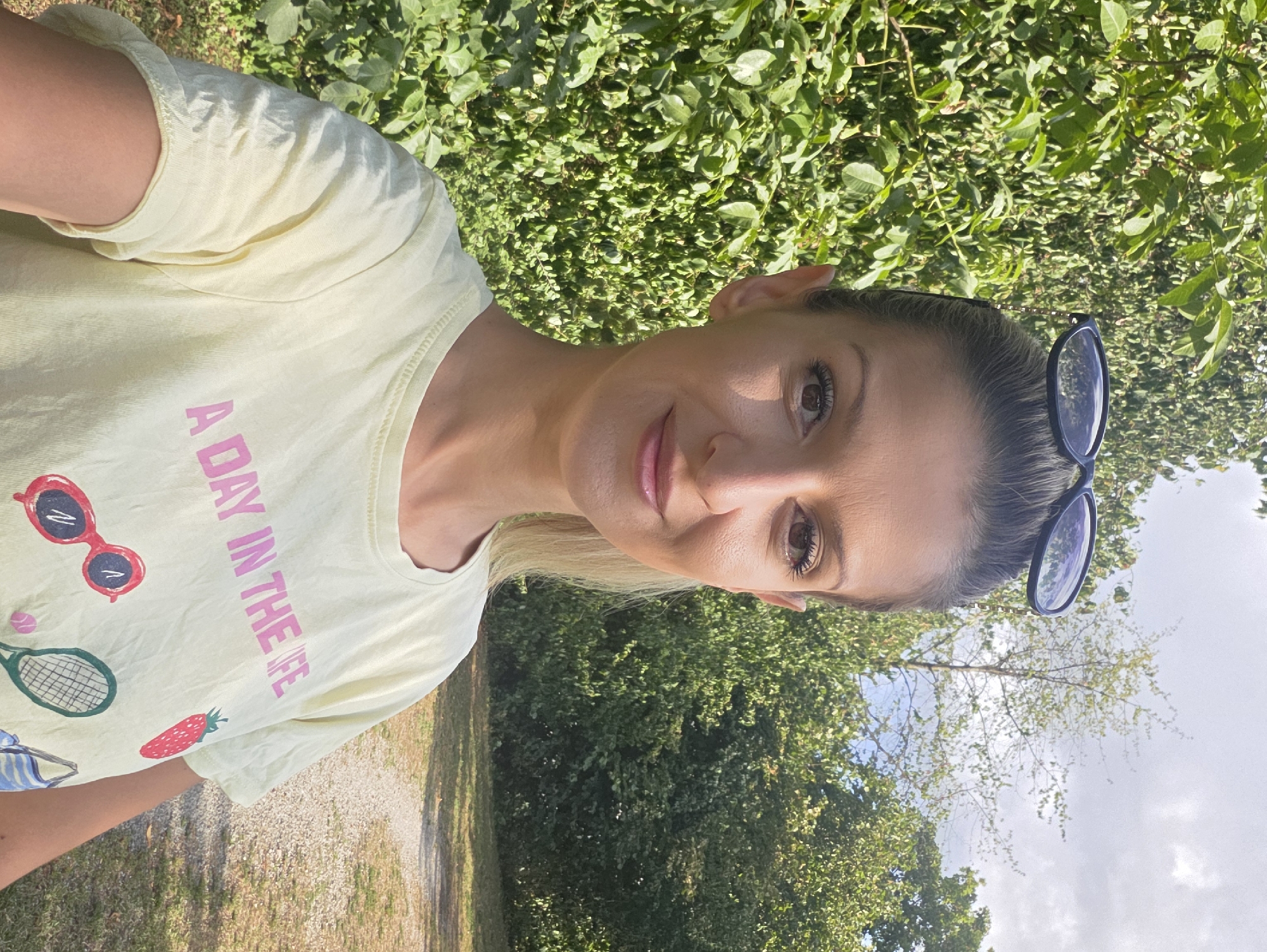 A woman is taking a selfie outdoors. She has blonde hair pulled back and is wearing sunglasses on top of her head. She is wearing a light yellow t-shirt with pink lettering that says "A DAY IN THE LIFE," and has cartoon drawings of a strawberry, sunglasses, and a tennis racket on it. She is standing in front of a dense green hedge, with trees and a path visible in the background. The sky is cloudy. The image does not provide specific details about the location within the city of Munich, Germany.