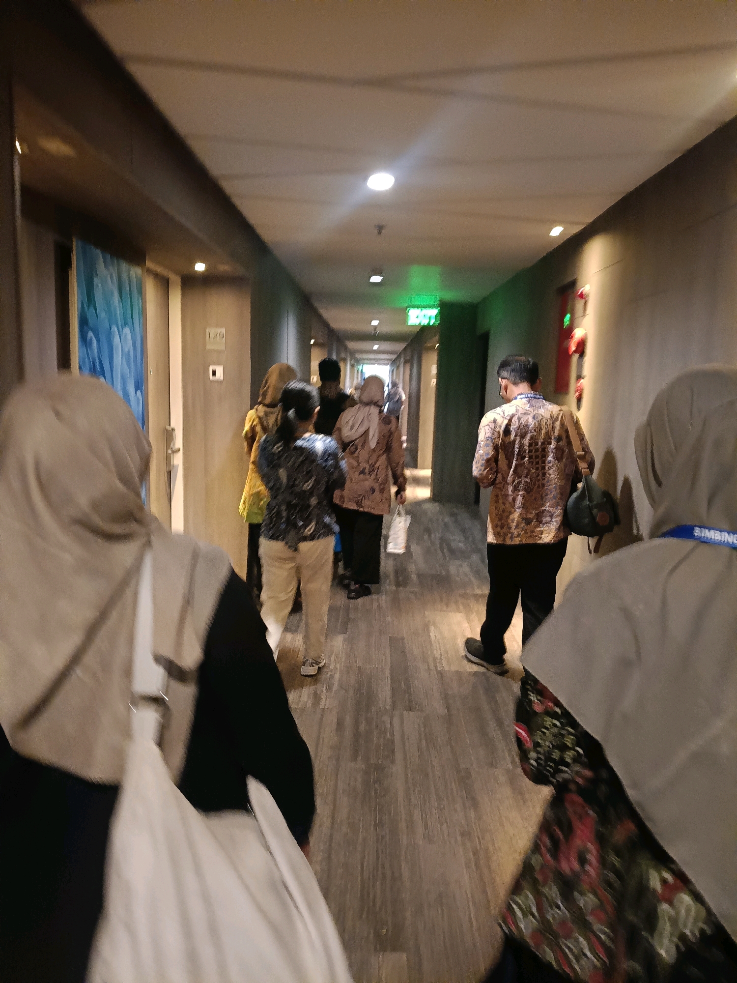 The image depicts a hallway inside a building, possibly a hotel or conference center. Several people are walking down the hallway, some from the back and others from the side. The hallway has a wooden floor and gray walls. There is a piece of art on the left wall, and a sign that says "EXIT" illuminated in green on the right side, indicating an emergency exit. The individuals are dressed in casual to semi-formal attire, and some appear to be wearing identification badges. The context suggests a public or semi-public indoor space in North Jakarta, Indonesia.