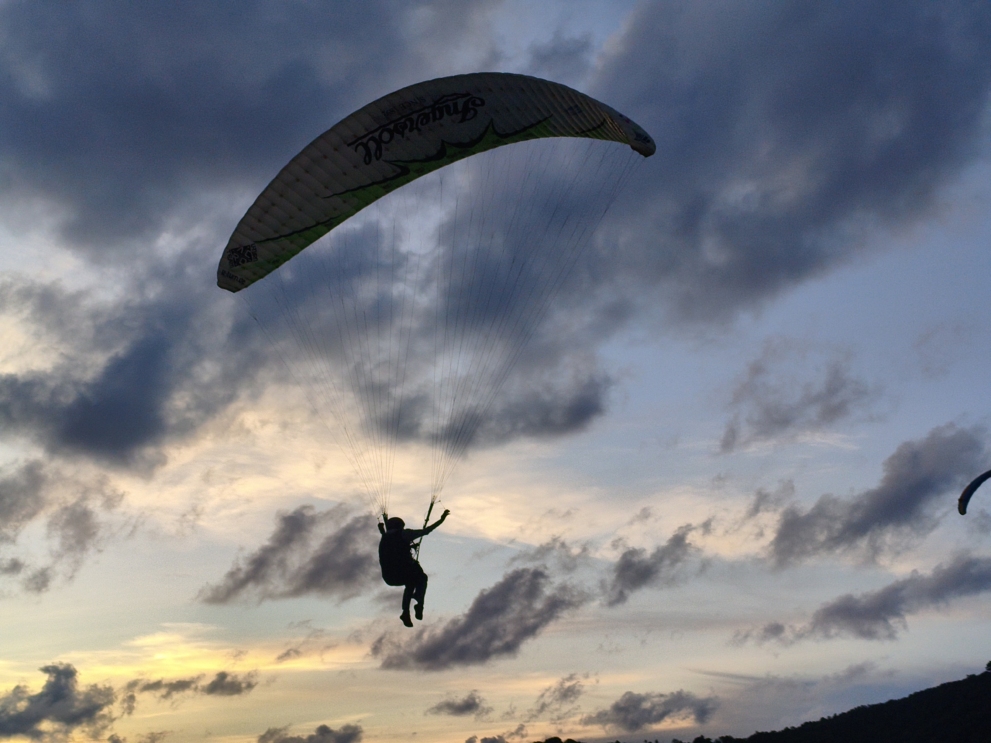 A paraglider is silhouetted against a twilight sky, with a paraglider canopy visible in the upper portion of the frame. The paraglider is in a seated position, suspended by lines from the canopy. The sky is filled with dramatic clouds, with patches of orange and yellow light suggesting the last rays of the sun. In the lower right corner, the dark silhouette of a hilly coastline is visible. The overall scene conveys a sense of freedom and adventure.
