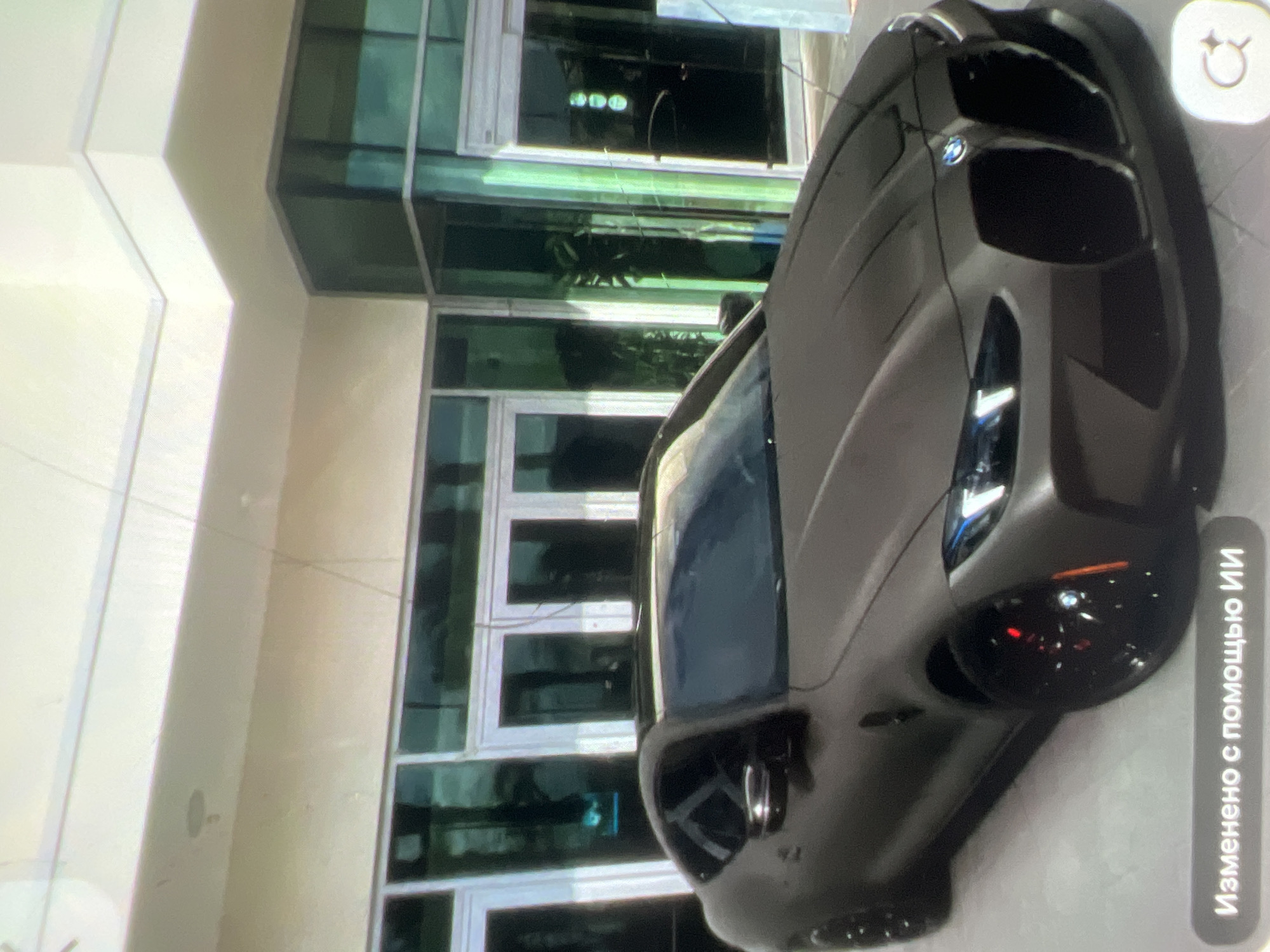 A matte brown coupe is parked in front of a modern building with large glass doors and windows. The car's headlights are illuminated. In the bottom left corner, a gray banner reads "Изменено с помощью ИИ" (Edited with AI). In the bottom right corner, there is a white circular icon with a magnifying glass.