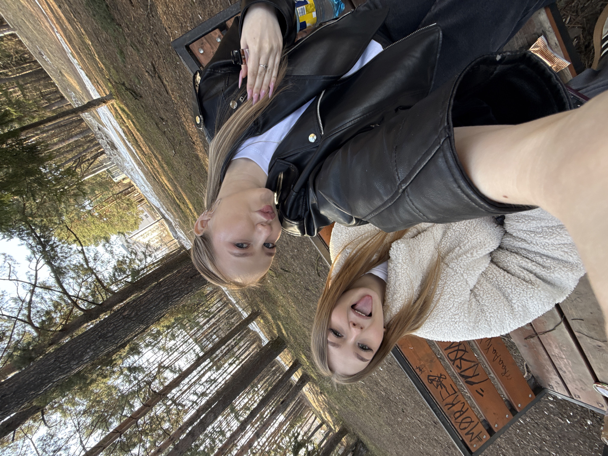 Two young women are sitting on a wooden bench in an outdoor environment. The bench has graffiti on it. One woman is wearing a black leather jacket over a white t-shirt, while the other is wearing a light-colored, fluffy jacket. The background consists of trees and possibly a body of water. Based on the presence of the city name "Chaikovsky" in the graffiti on the bench, it is likely that the location is in Chaikovsky, Russia.