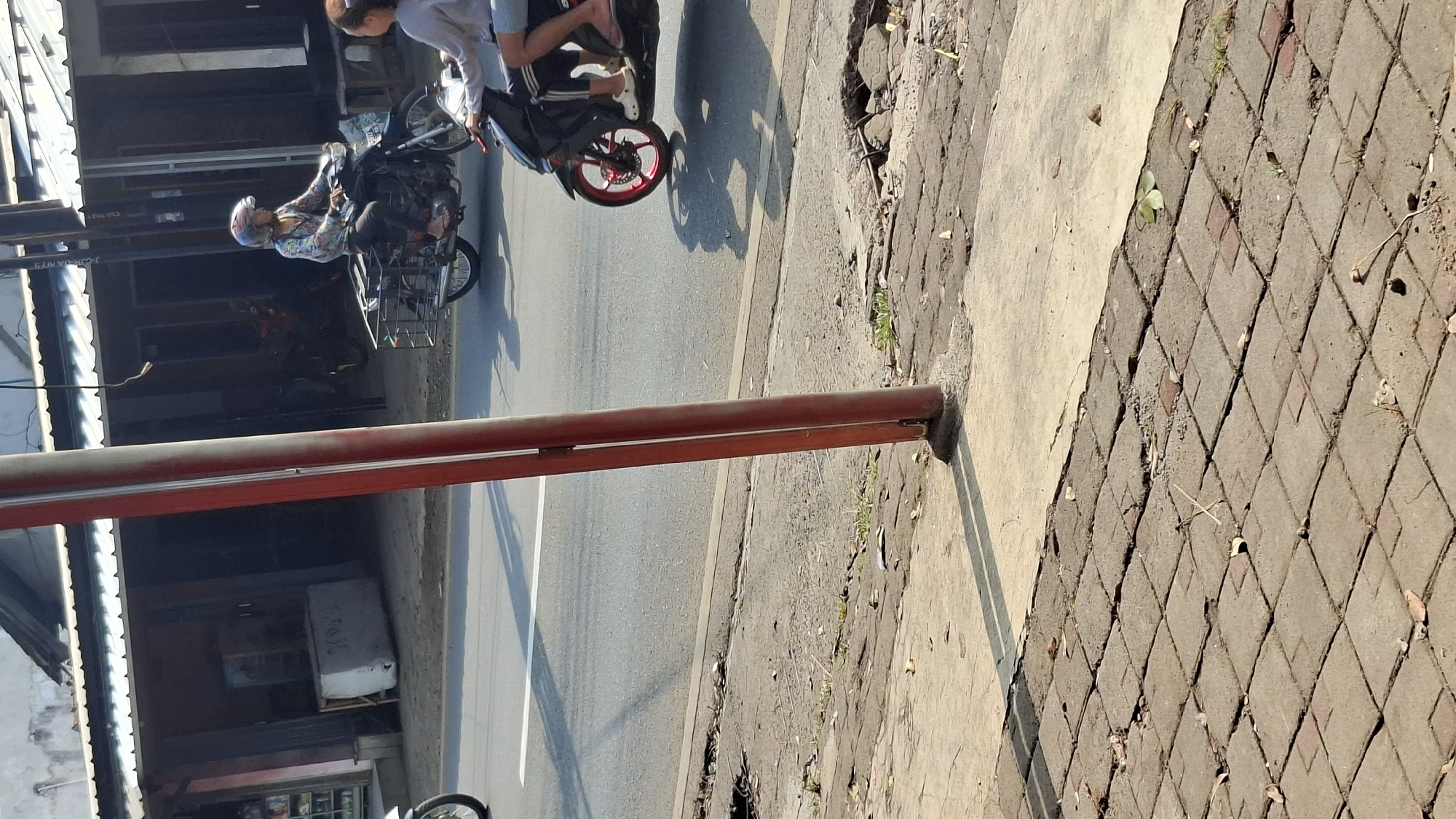 In Cilaku, Indonesia, two motorbikes are depicted on a paved road in daylight. The first motorbike, on the left, is ridden by an individual wearing a helmet and a patterned shirt. This motorbike has a metal basket attached to its rear. The second motorbike, on the right, carries two individuals. The rider is partially visible, and the passenger, a female wearing a gray top and white shoes, is looking to the left. The road surface shows signs of wear and tear, with visible cracks and an uneven texture. Adjacent to the road is a sidewalk paved with interlocking rectangular bricks, some of which appear damaged or displaced. A reddish-brown pole stands prominently in the foreground, casting a distinct shadow on the sidewalk. In the background, buildings with corrugated roofs line the street, suggesting a commercial or residential area. Sunlight creates strong shadows, indicating a sunny day.