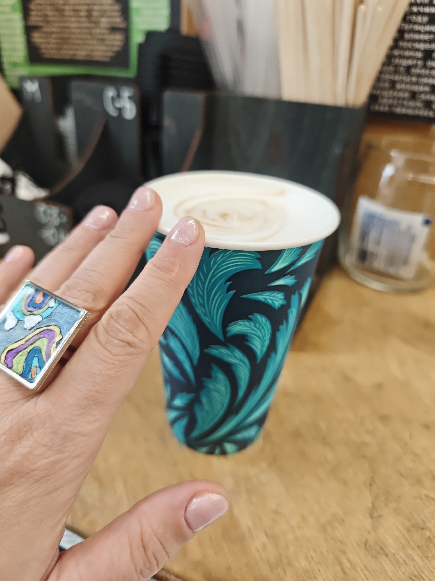 A photograph depicts a hand wearing a large, rectangular ring with inlaid, colorful abstract designs. The hand is positioned near a takeaway coffee cup adorned with a teal and black leafy pattern. The cup rests on a wooden surface. In the background, out-of-focus details suggest a cafe setting with organizational containers for supplies, possibly in Moscow, Russia.