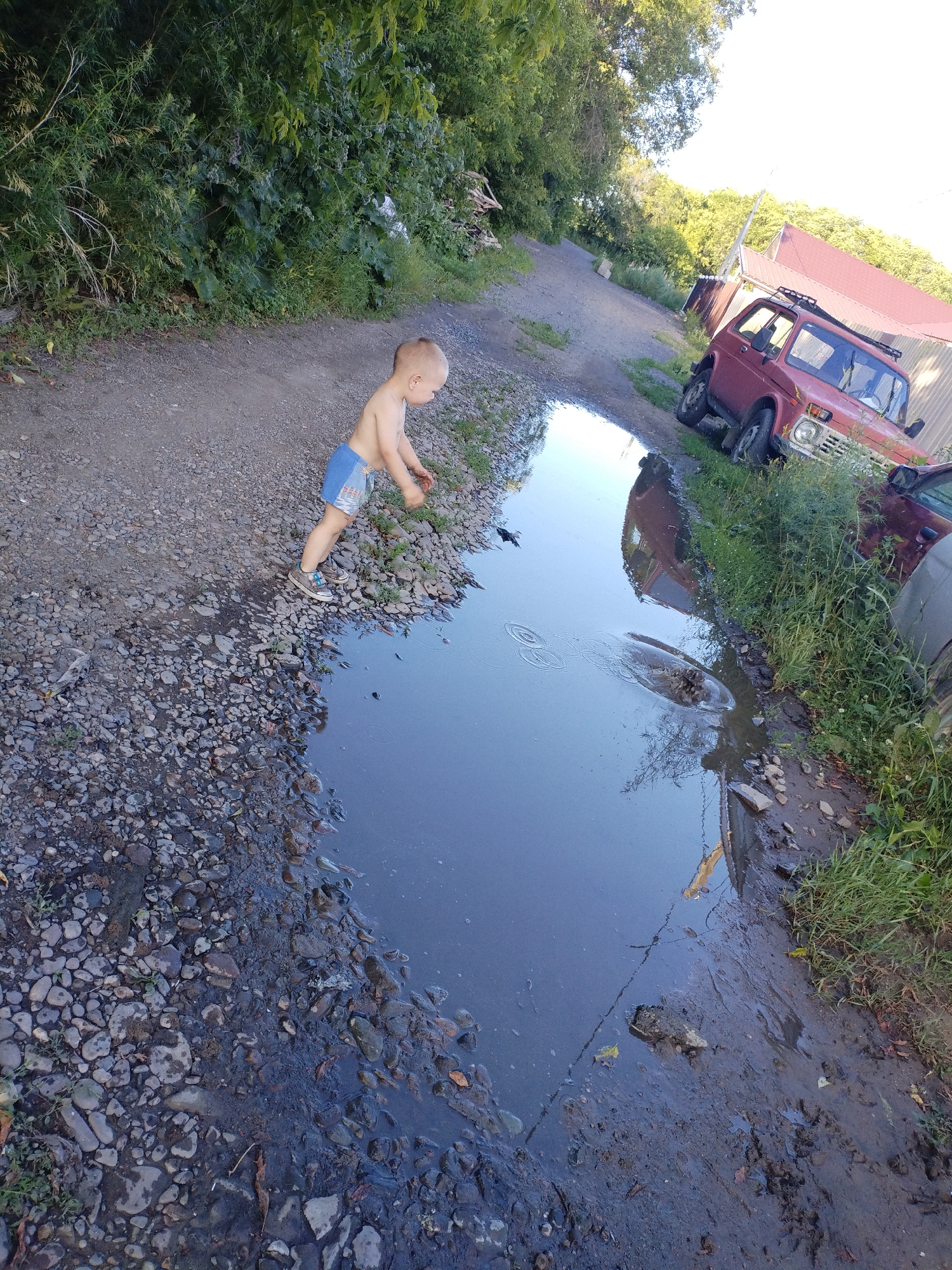 A young, shirtless child wearing blue shorts and grey sneakers stands by a large muddy puddle on a gravel path. The child appears to be throwing something into the puddle. The background consists of lush green foliage and trees, a red four-wheel-drive vehicle with a roof rack, and a corrugated metal building with a red roof. The scene is set in an outdoor, possibly rural or semi-urban area within the city of Komsomolsk-on-Amur, Russia. The lighting suggests it is daytime, with sunlight illuminating the scene.
