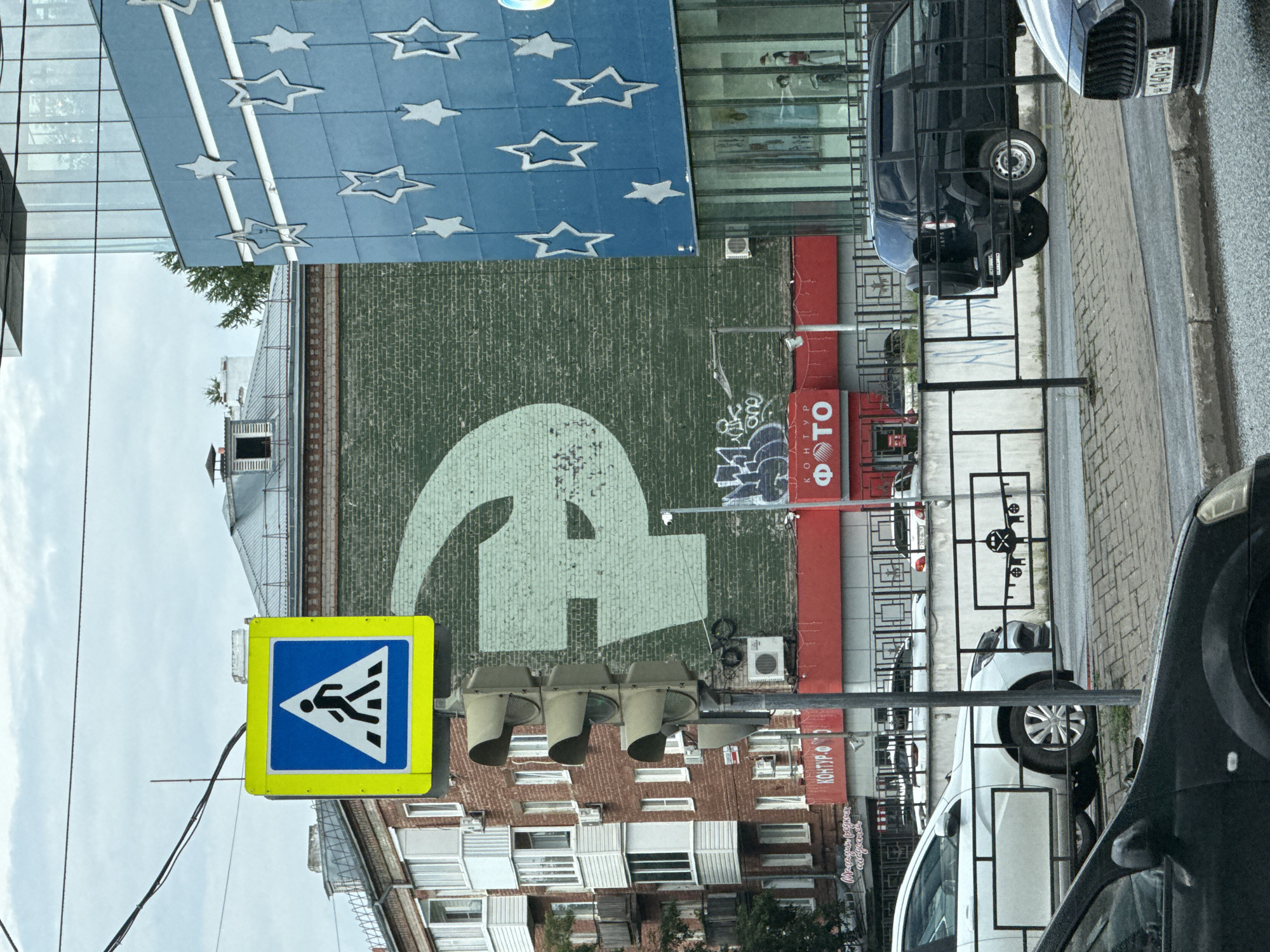 The image depicts a street scene in Izhevsk, Russia, under an overcast sky. A prominent feature is a large, pale green hammer and sickle symbol painted on the brick wall of a building. In the foreground, a traffic light with three signal heads stands on a pole, next to a yellow and blue pedestrian crossing sign. Adjacent to the brick building, a modern blue building with white star decorations is visible. A red awning with the word "ФОТО" (photo) in white letters stretches across the lower part of the brick building. A black SUV and several white cars are parked or driving on the street, separated from the sidewalk by a black metal fence. The sidewalk is paved with cobblestones.