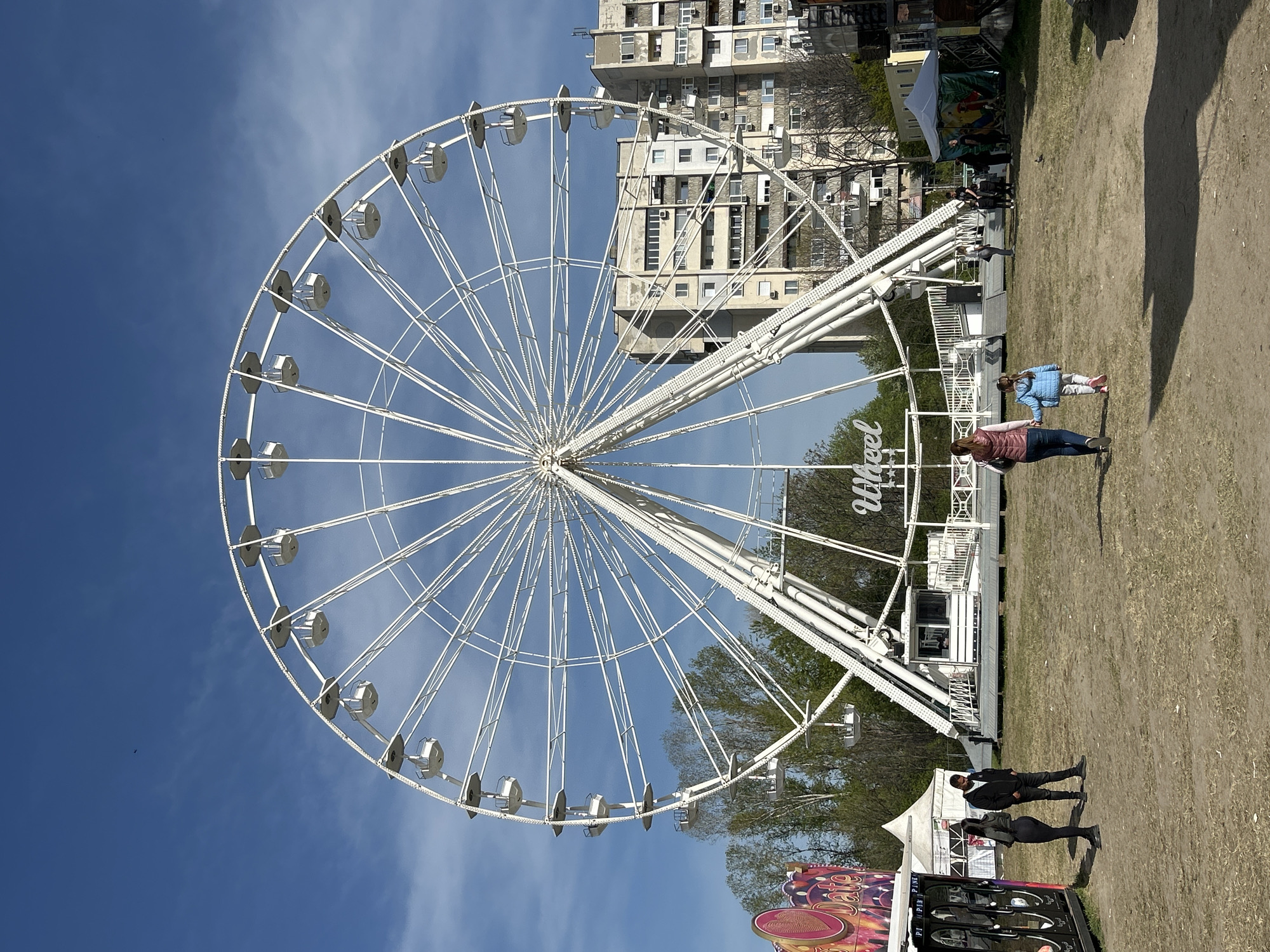 This image depicts a vibrant outdoor scene dominated by a large, white Ferris wheel positioned on the left side of the frame. The Ferris wheel is intricately designed with numerous spokes and passenger gondolas, and the word "Wheel" is visible on its support structure.
The background features a clear, bright blue sky with scattered, wispy clouds. Behind the Ferris wheel, several multi-story residential buildings with numerous windows and air conditioning units are visible, indicating an urban setting. Sparse trees are also present between the structures and the Ferris wheel.
The foreground and mid-ground consist of dry, earthy ground, suggesting an open field or fairground. Several individuals are present, walking across the scene. Towards the center-right, two women and a child are seen. One woman wears a brown/maroon jacket, another a blue jacket, and the child is also in a blue jacket. Near the bottom-right corner, two adults in dark attire stand near a white tent. Other people can be faintly seen closer to the base of the Ferris wheel and other distant structures.
The activity taking place appears to be a public event, likely a fair or carnival, given the presence of the Ferris wheel and tents. A colorful booth with partial text, including "PIPI" and what might be "Late" or similar, is visible at the very bottom-right. The strong sunlight and clear skies indicate a bright day, likely midday or early afternoon. The overall setting suggests a temporary amusement event within a city, specifically Plovdiv, Bulgaria.