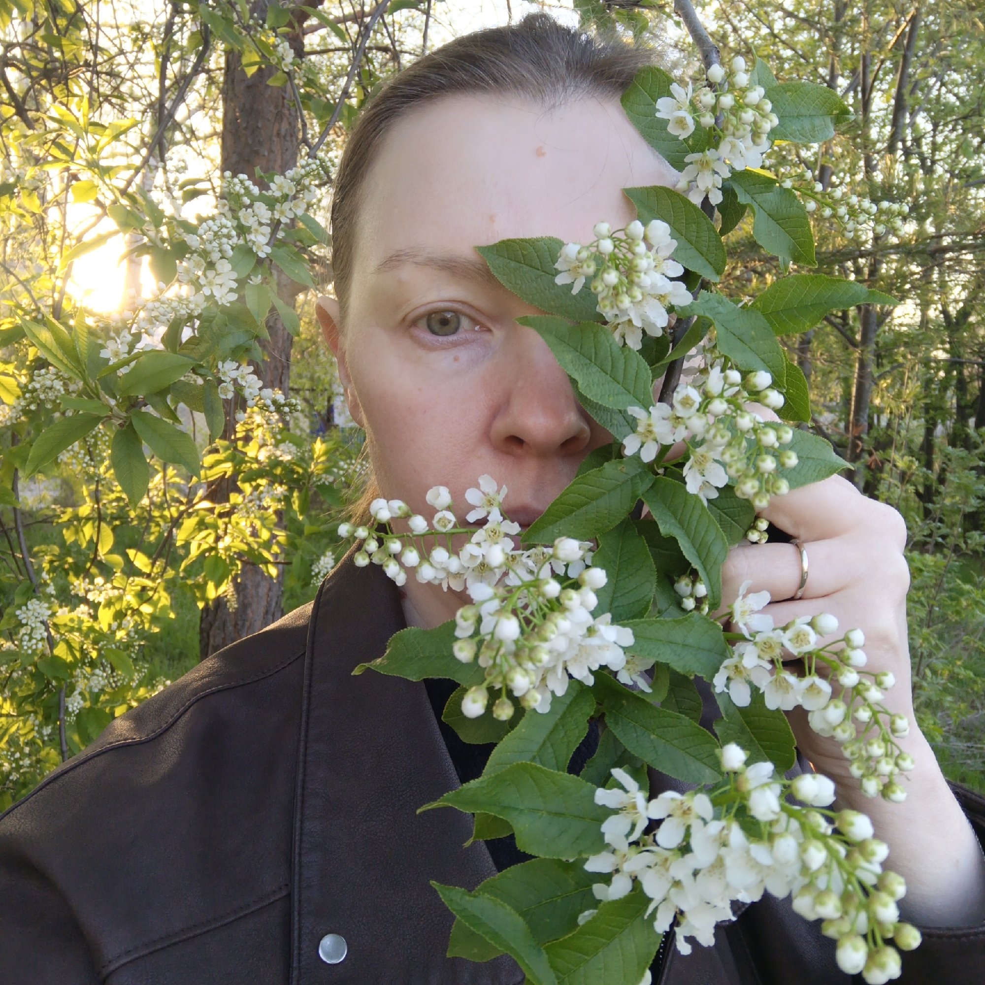 A person wearing a brown leather jacket is holding a branch of flowering white blossoms in front of their face. The person's left eye is visible, and a gold ring is on a finger of their right hand, which holds the branch. The background consists of trees and foliage, with sunlight filtering through the branches. The location is Chaikovsky, Russia.