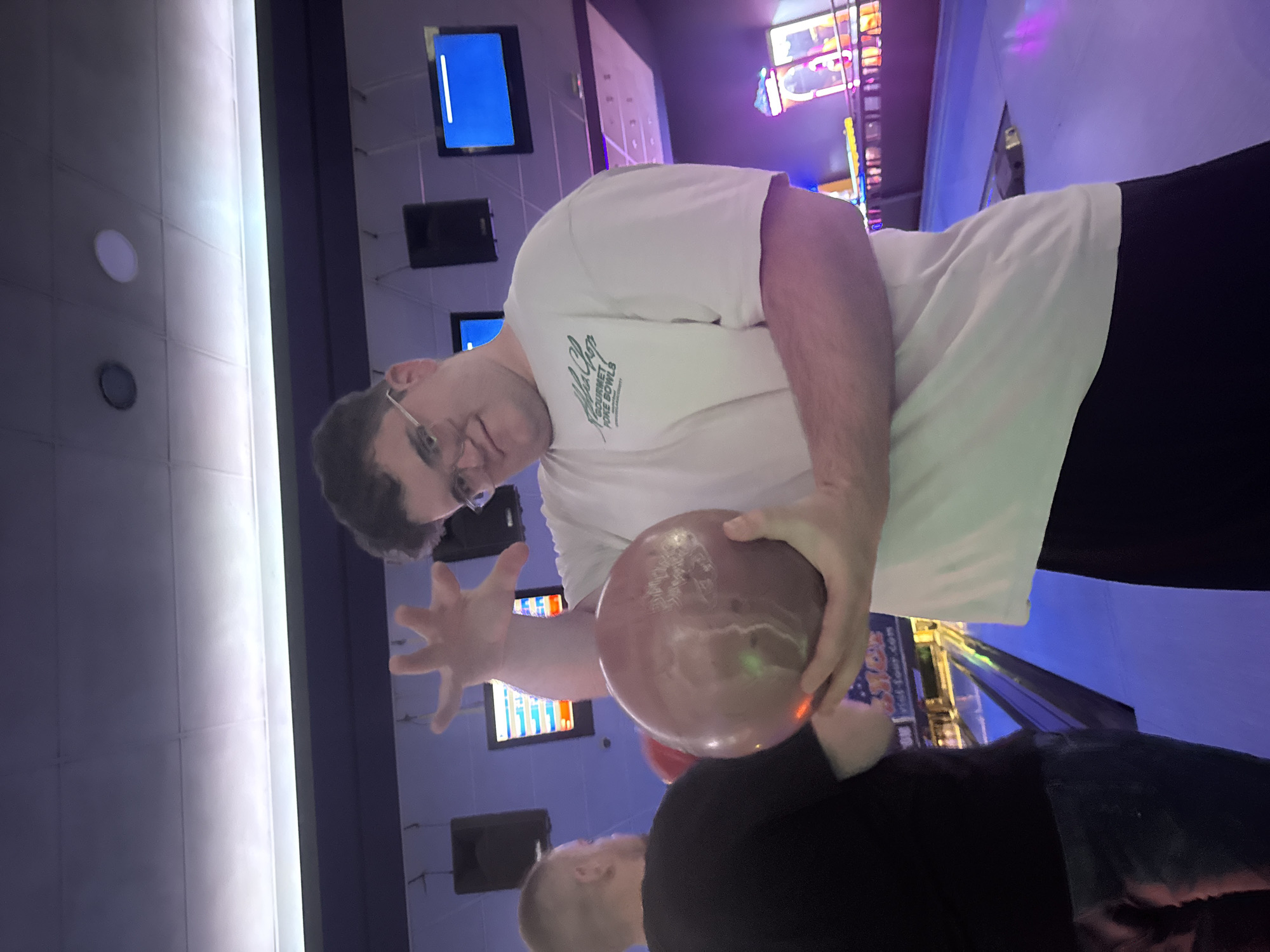 A man wearing glasses and a white t-shirt with text on it is holding a bowling ball in a bowling alley. Another person with short hair, wearing a black shirt is standing next to him. The alley has screens and lights. The location is unknown and may not be in Pleven, Bulgaria.