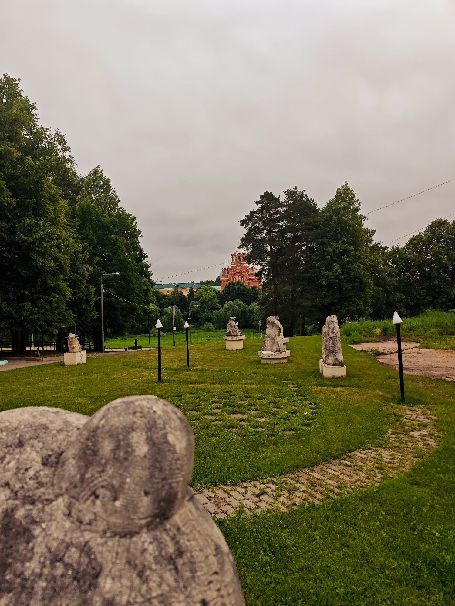 The image depicts a park-like setting with several stone sculptures placed on a grassy lawn. In the background, a large, red-brick church with white domes and gold accents is visible. The sky is overcast, and the surrounding area is filled with lush green trees. A paved path winds through the grass, and there are several lampposts scattered throughout the scene. The foreground features a close-up of a blurred stone sculpture, suggesting it is a significant element of the park's display. The overall atmosphere is serene and somewhat somber due to the cloudy weather. This scene is located in Khotkovo, Russia, likely at the Museum-Estate Abramtsevo, a renowned art museum and cultural heritage site.