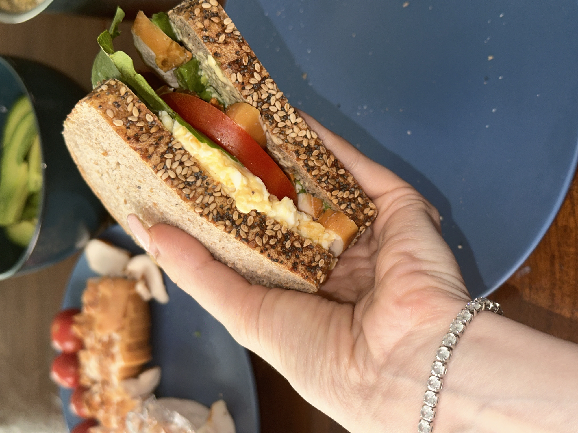 A close-up captures a hand holding a sandwich. The sandwich is made with multi-grain bread, and the filling appears to include egg, lettuce, tomato, and what may be a spread or another filling. The hand is wearing a silver bracelet. Beneath the sandwich and hand, a blue plate is partially visible, containing sliced avocados in the top-left corner, and a salad with what seems like cherry tomatoes, croutons, and lettuce on the bottom. A wooden table is visible to the right of the plate. The location is unknown.