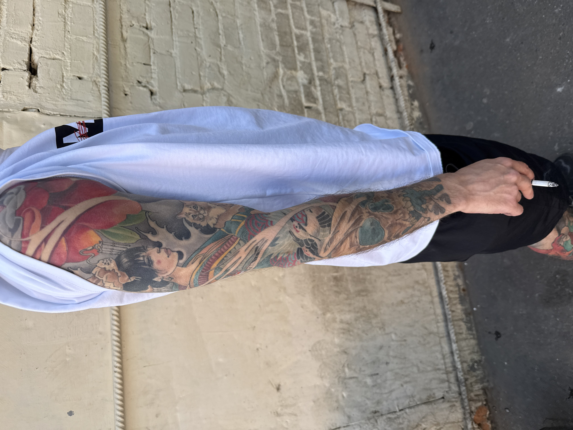 A person stands outdoors in what appears to be an urban area of Moscow, Russia, positioned against a light-colored brick wall with a textured horizontal molding. The individual, seen from the right side, wears a white short-sleeved t-shirt with a black and red logo on the upper right sleeve, and dark-colored shorts. Their right arm is heavily tattooed with a full sleeve in a Japanese-inspired style, featuring a female figure with black hair and an ornate hairpiece, vibrant red and orange flowers (likely peonies), a small cat, one or more cranes, and a green-blue skull on the lower forearm. The right hand is visible, holding a lit cigarette between the thumb and index finger. A colorful tattoo is partially visible on the lower right leg, just above the ankle. The ground is dark gray asphalt or concrete, showing some moisture and minor debris. The brick wall behind the person exhibits wear, cracks, and discoloration.