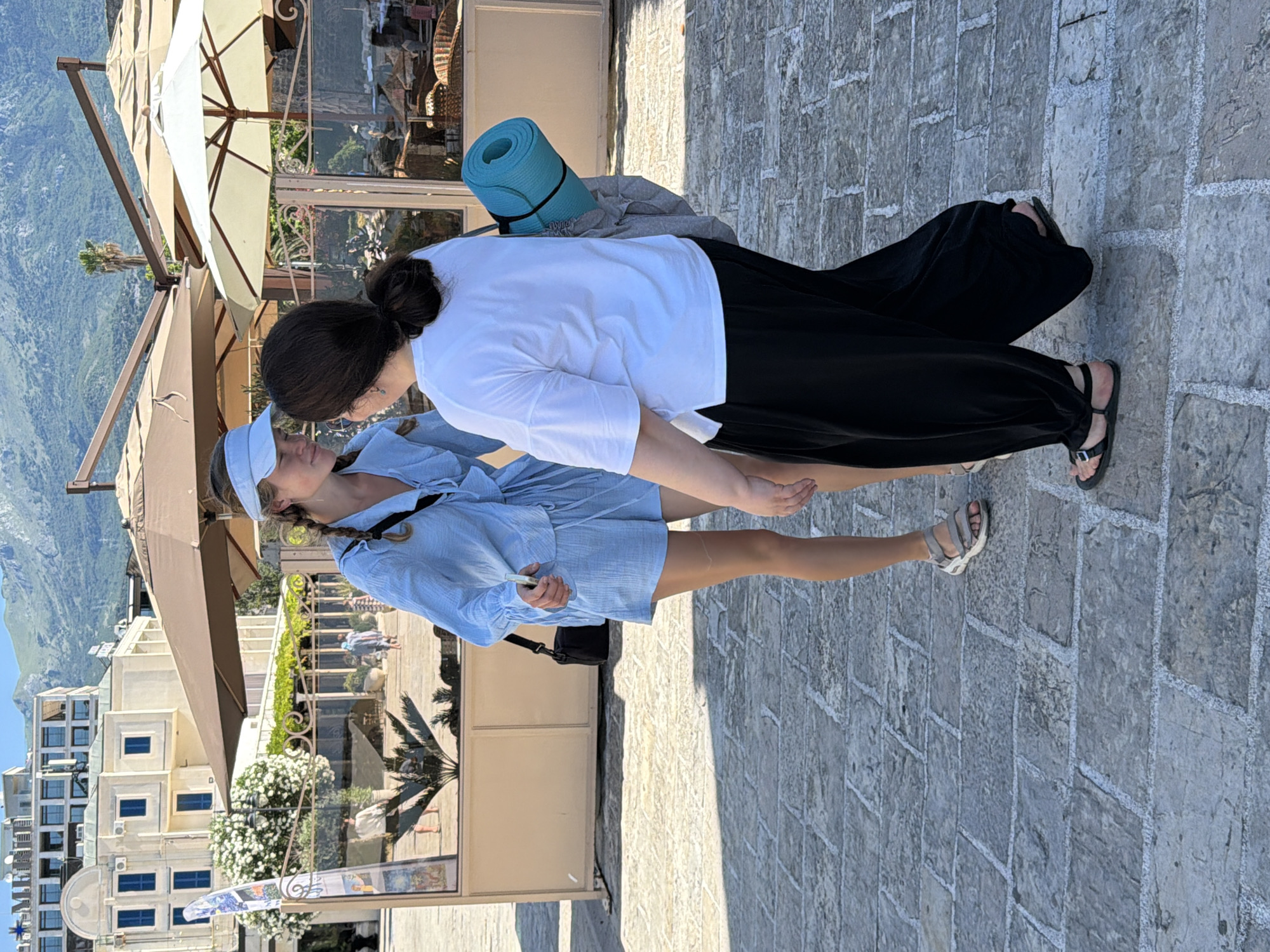 Two young women are engaged in conversation on a cobblestone street in Budva, Montenegro. The woman on the left, wearing a light blue collared shirt and matching shorts, has her hair in braids and is wearing a light blue visor. A black crossbody bag is slung over her shoulder. The woman on the right is wearing a white t-shirt and loose black pants. A rolled-up teal yoga mat is strapped to her back, along with a grey bag. In the background, there are buildings with white facades and blue windows, shaded outdoor seating areas with umbrellas, and a backdrop of lush green mountains. The overall atmosphere appears to be a sunny day in a coastal city.