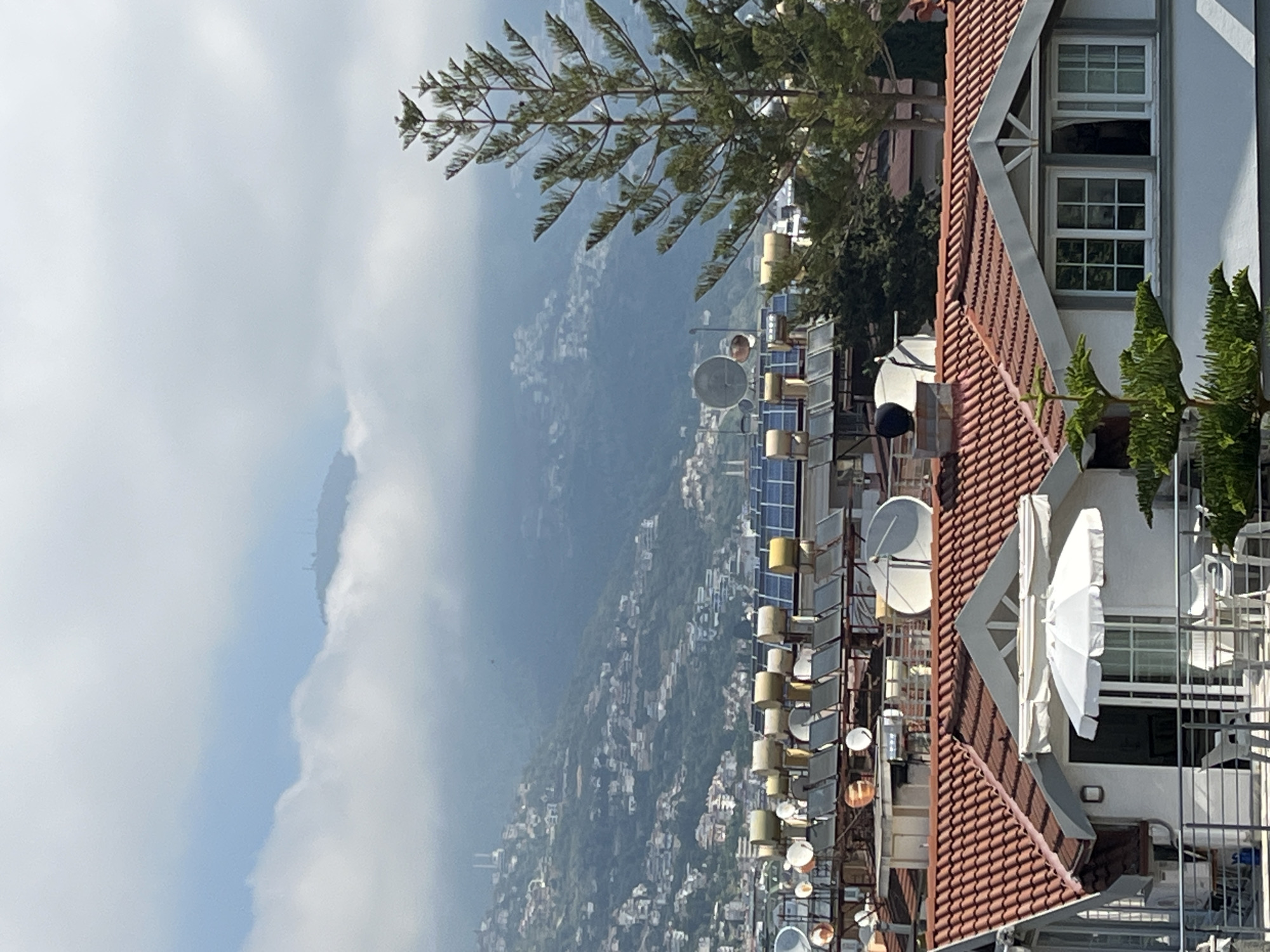 The image presents a panoramic view of a city nestled on a hillside, extending towards distant hazy mountains, under a partly cloudy sky. The perspective is from a high vantage point, looking down upon the scene.
In the foreground, on the right side of the image, is a building featuring a prominent red tiled roof. A large, open white umbrella is visible on a balcony or terrace section of this building, alongside white-framed windows. Green foliage from trees and shrubs is scattered around this structure.
The midground reveals a dense collection of various communication devices, including multiple satellite dishes and an array of solar panels. These are mounted on what appears to be a rooftop or a dedicated infrastructure, suggesting a hub of technology or energy generation.
Further into the background, the city's urban sprawl covers a vast hillside, with numerous buildings extending down towards the horizon. Beyond the city, large mountains rise, partially obscured by atmospheric haze and soft white clouds that dot the sky. The sky is a blend of light blue and white cloud cover.
The scene is captured during bright daytime, with no specific activity taking place. There is no visible text. The architectural style, combined with the mountainous coastal landscape, is consistent with the provided location context of Alanya, Turkey.